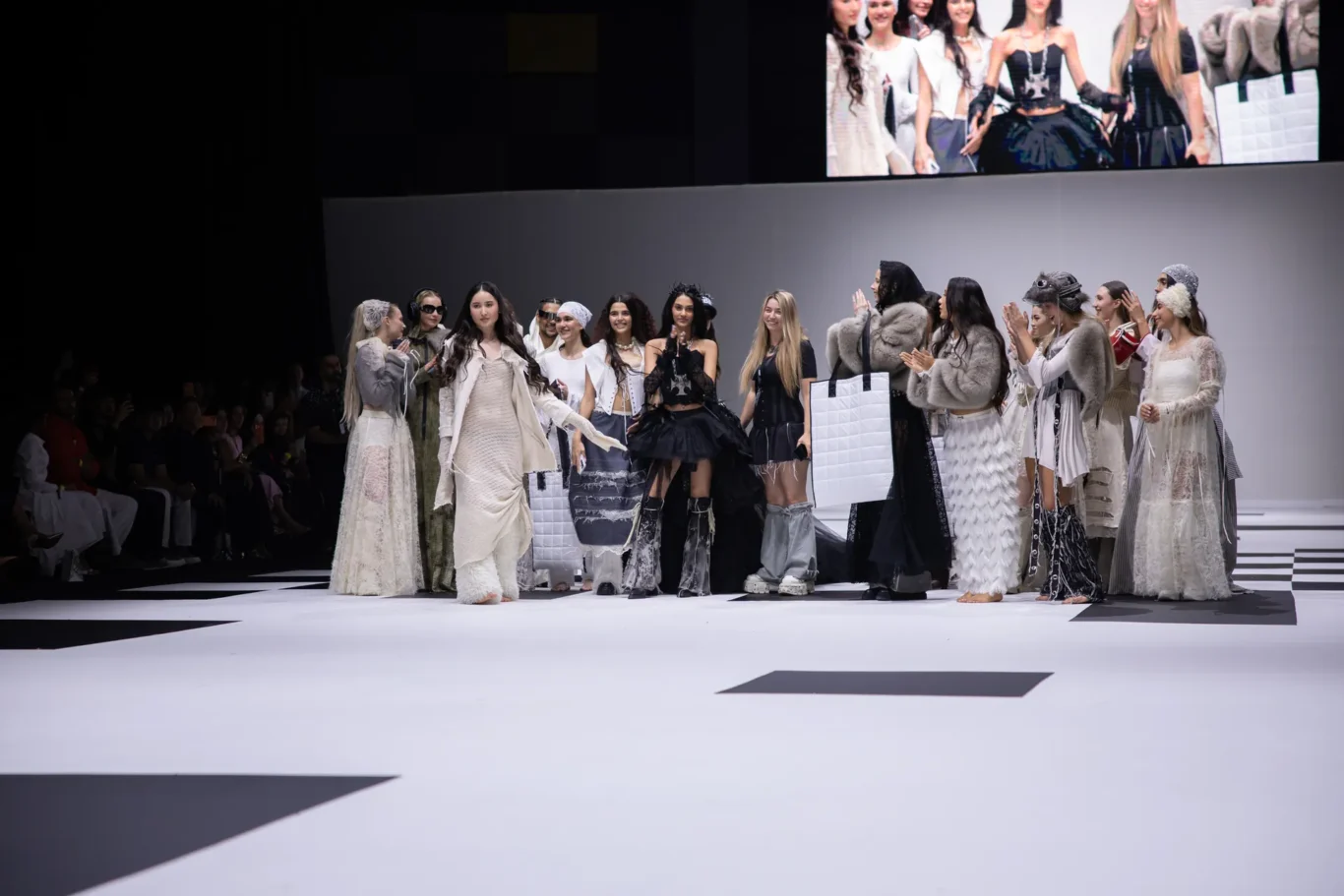 Models in glamorous monochrome outfits walk a runway during a fashion show, with an audience and a large screen above.