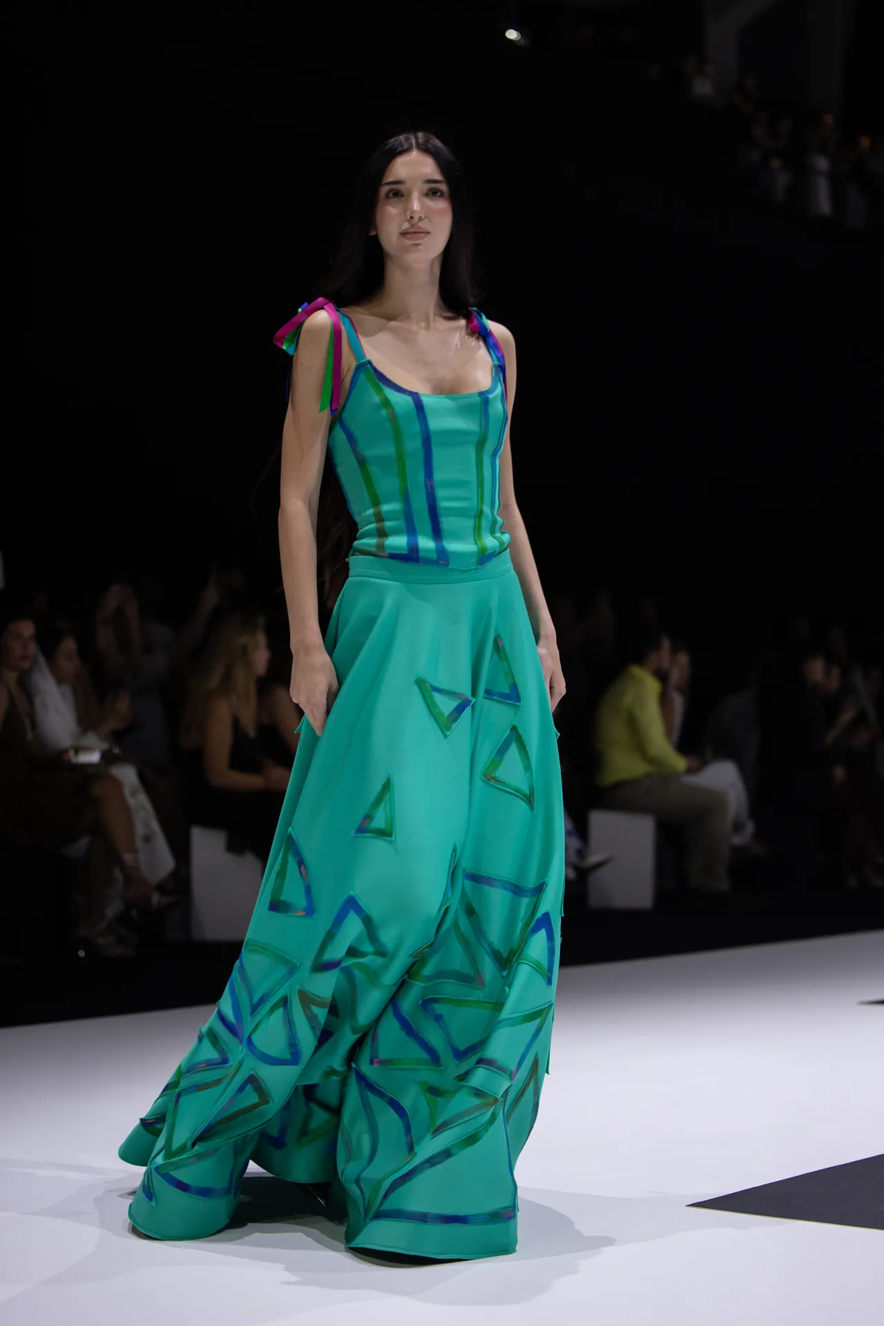 Female model walks a runway in a turquoise gown featuring blue-green triangle patterns.