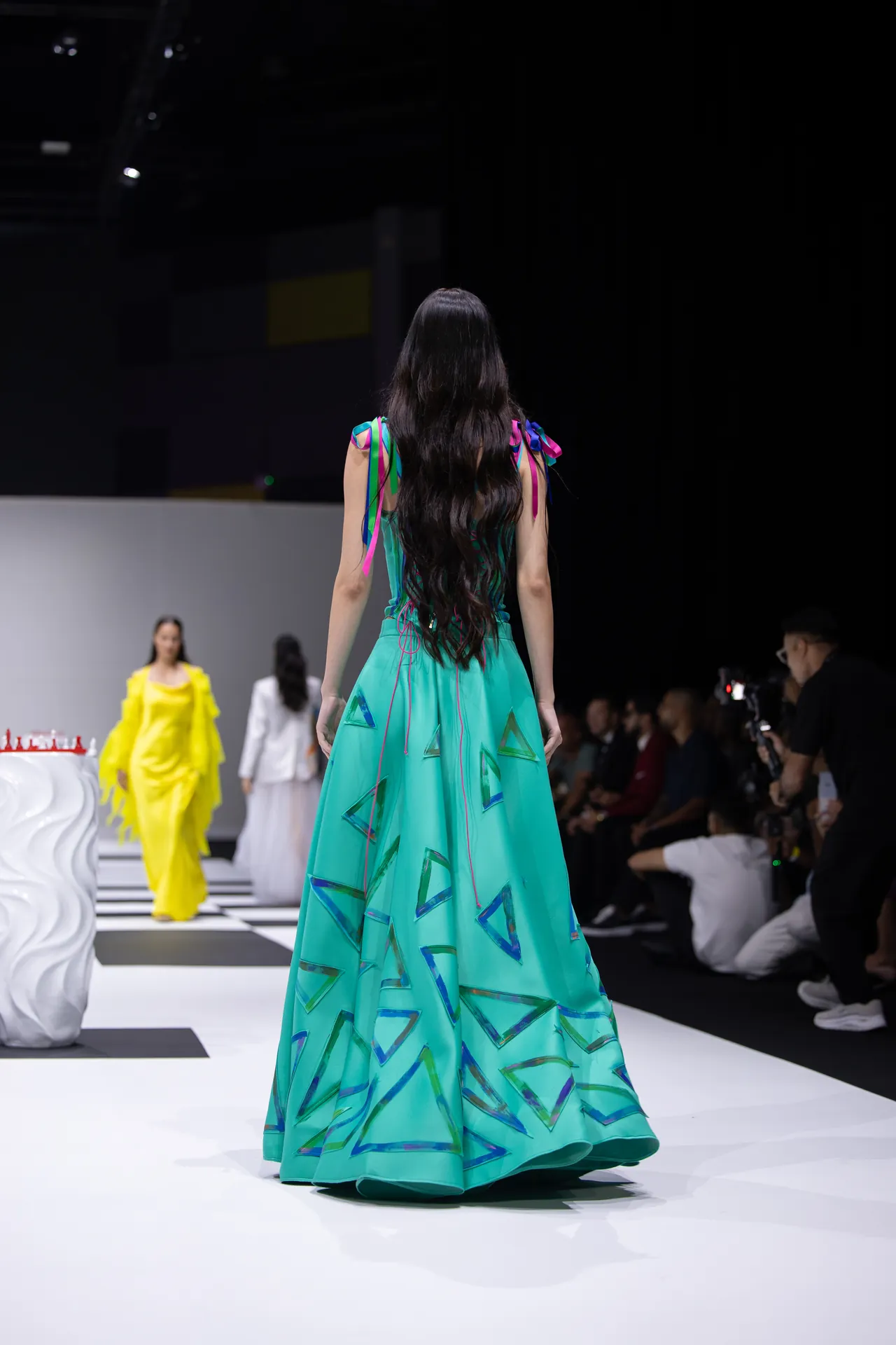 Back view of a model in a turquoise gown with blue-green triangle patterns walking the runway, audience in darkness beyond.