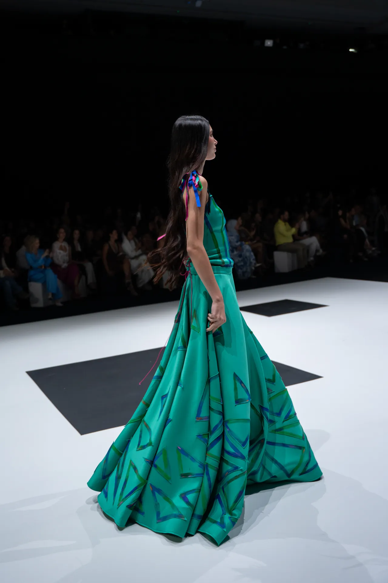 Side view of a female model walking the runway in a turquoise gown with geometric blue patterns, audience in shadow along the catwalk behind her.