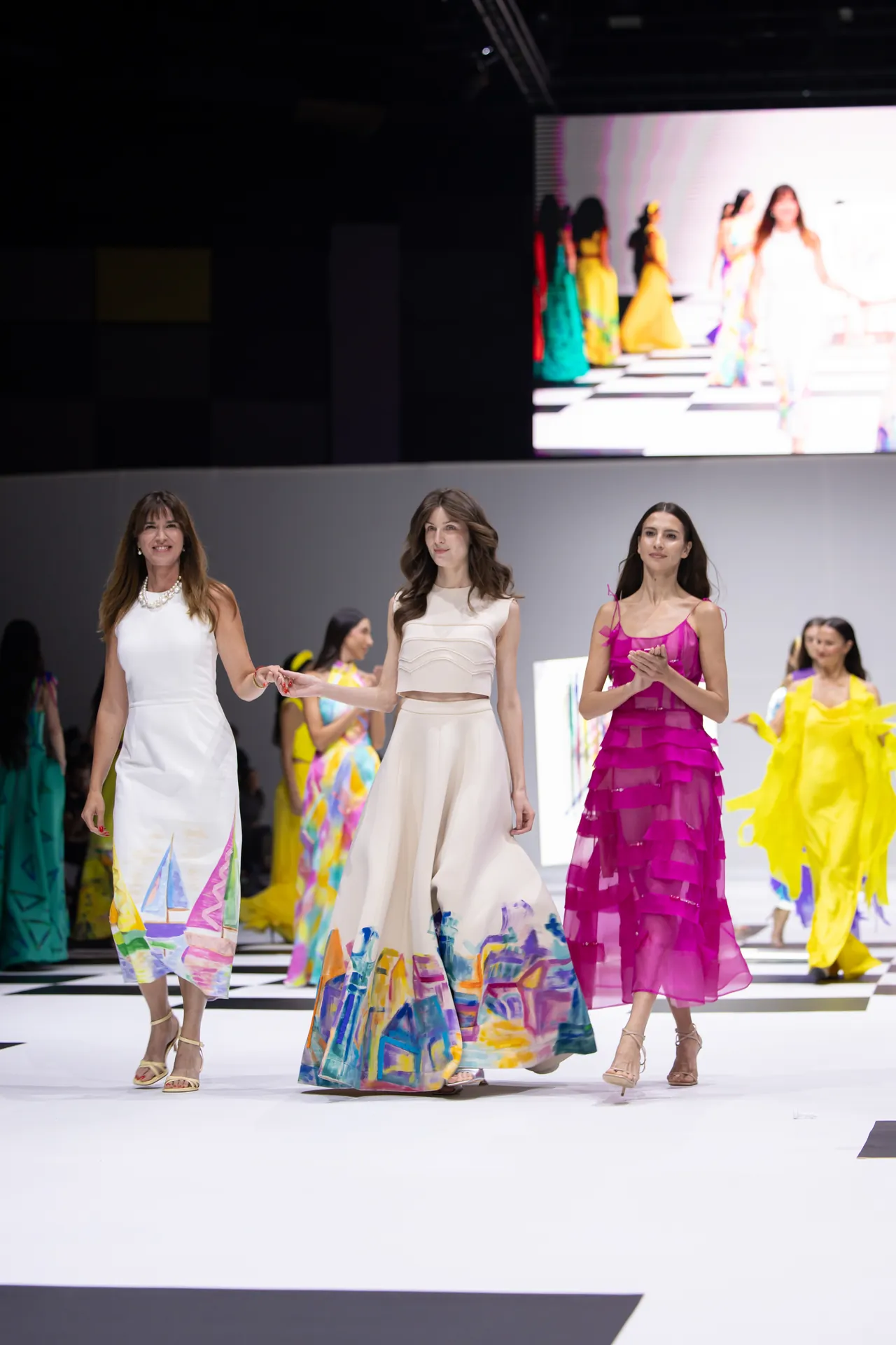 Three models walk a fashion runway in vivid evening gowns: white, cream, and magenta with colorful hem designs on a checkered floor.