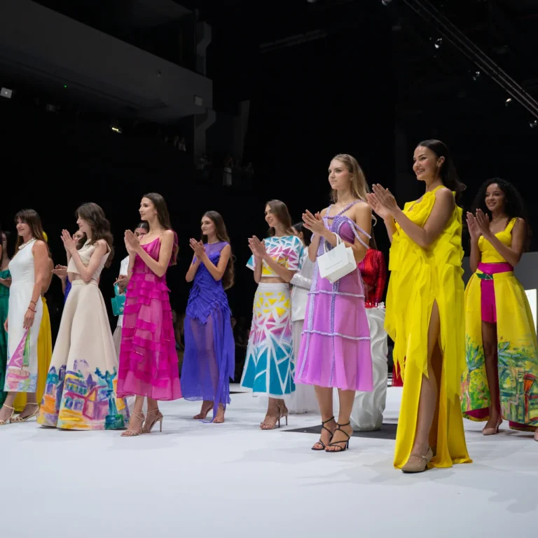 Group of models standing on a runway in bright, colorful dresses, applauding as the audience watches.
