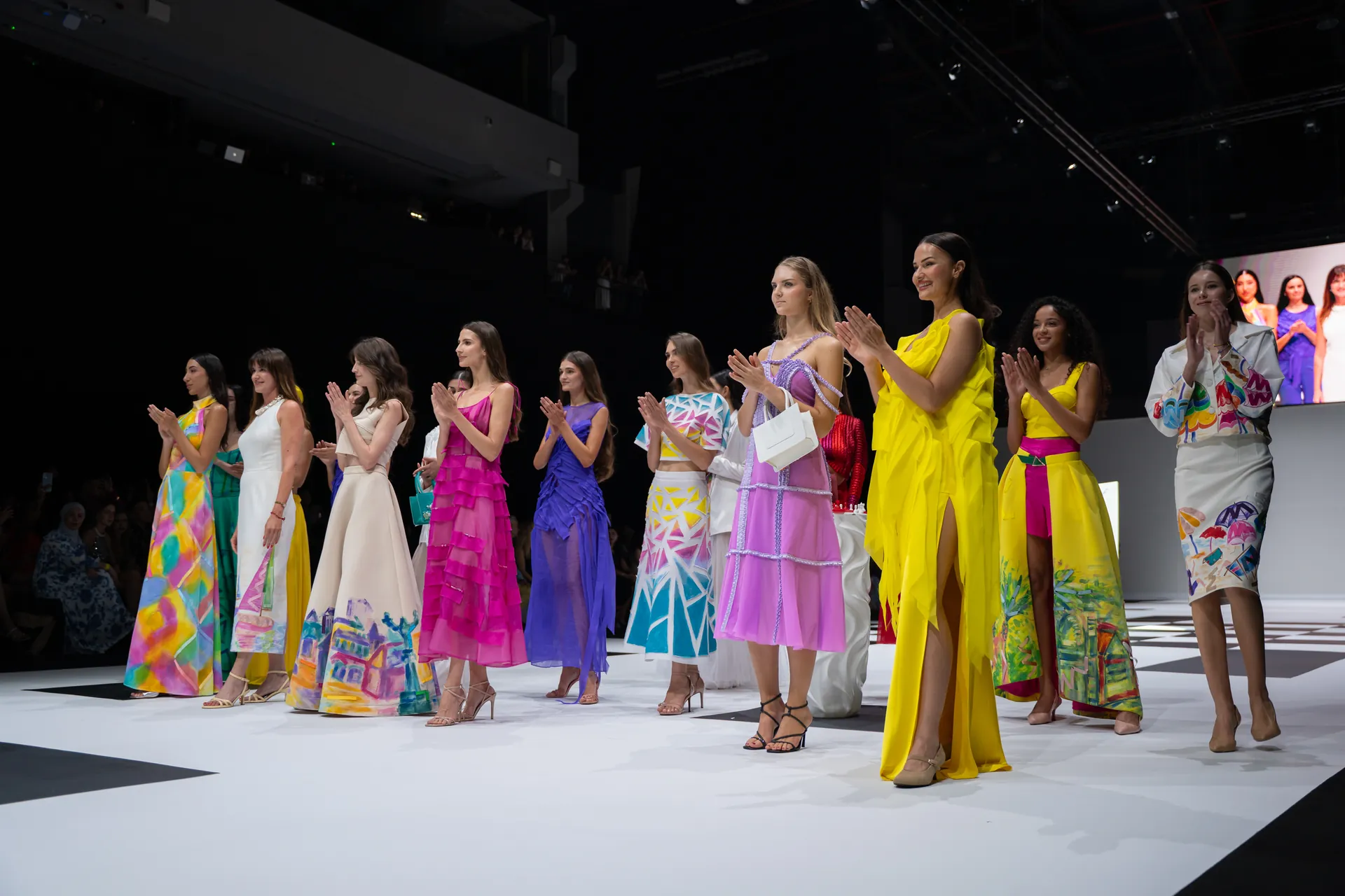 Group of models standing on a runway in bright, colorful dresses, applauding as the audience watches.