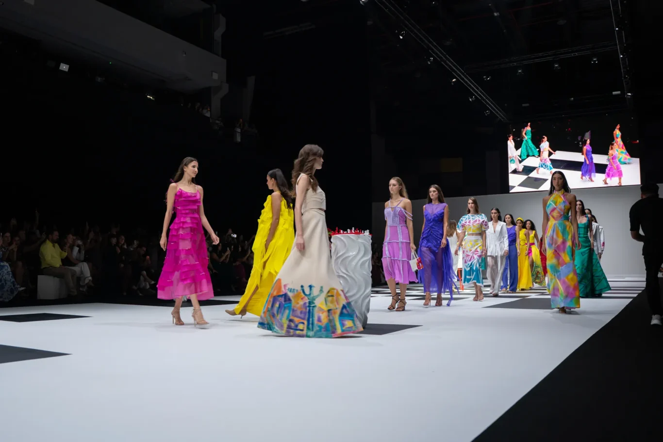 Models in colorful gowns walk a checkerboard runway at a fashion show, audience visible on the left.