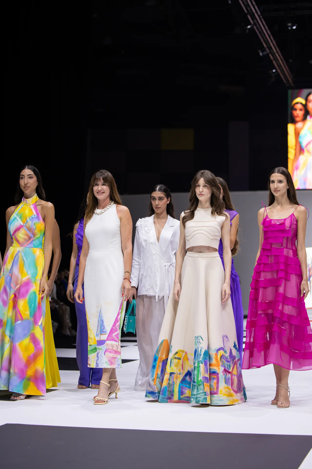 Models walk a fashion runway wearing vibrant gowns in pastel and bright colors.