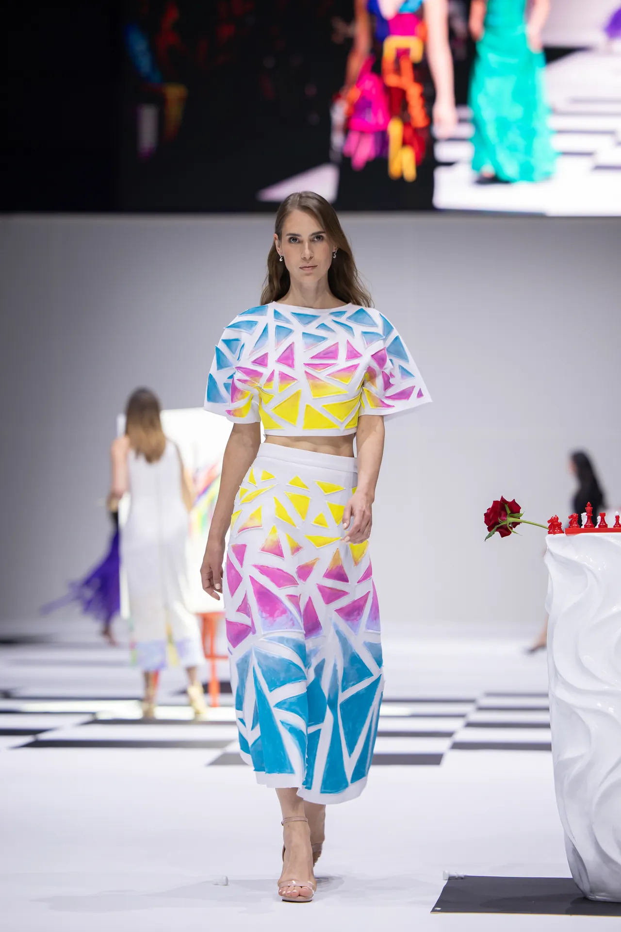 Female model walks the runway in a white two-piece outfit with blue, pink, and yellow geometric triangle patterns on a checkered floor.