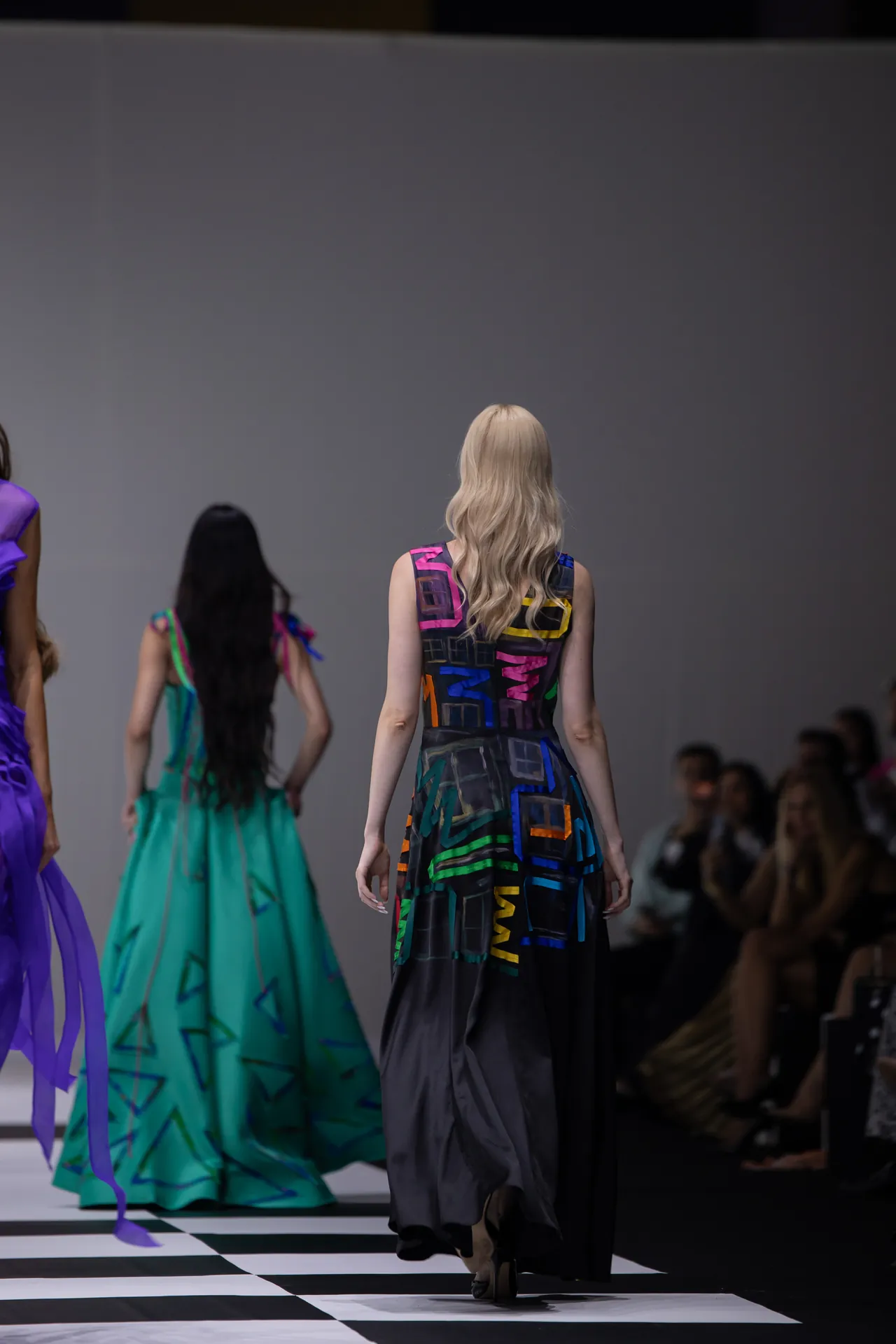 Model walking away on a fashion runway in a long black dress with a bright geometric print; audience visible on the sides against a plain backdrop.