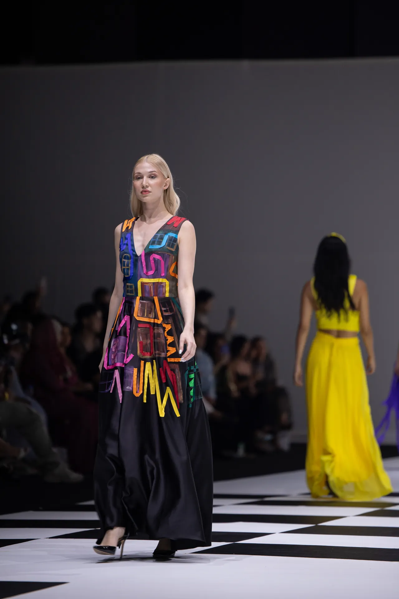 Model walks the runway in a sleeveless, multicolor geometric-print gown with high heels.