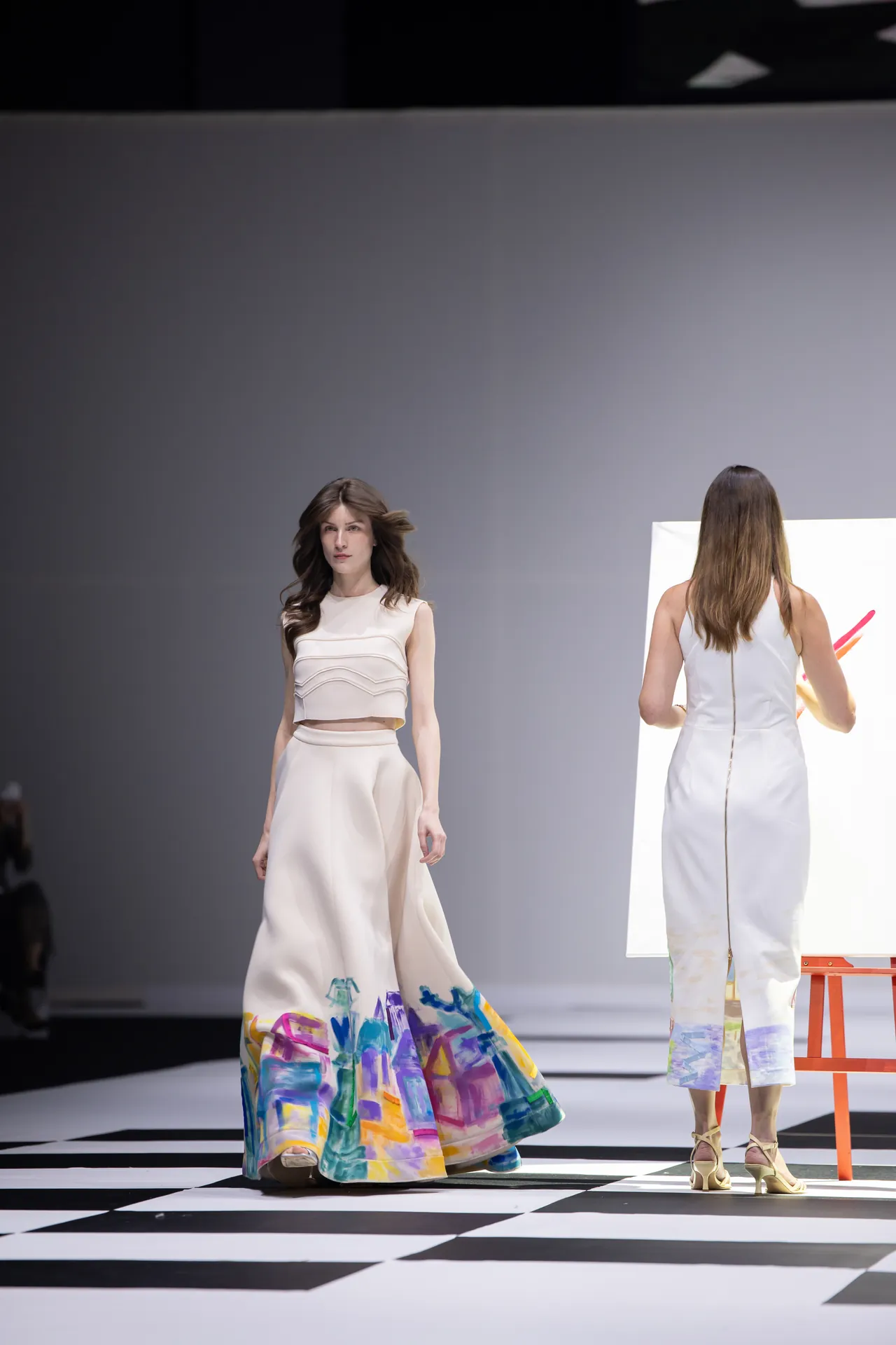 Model walks a runway in a pale cream crop top and full skirt with a colorful painted hem at a fashion show.
