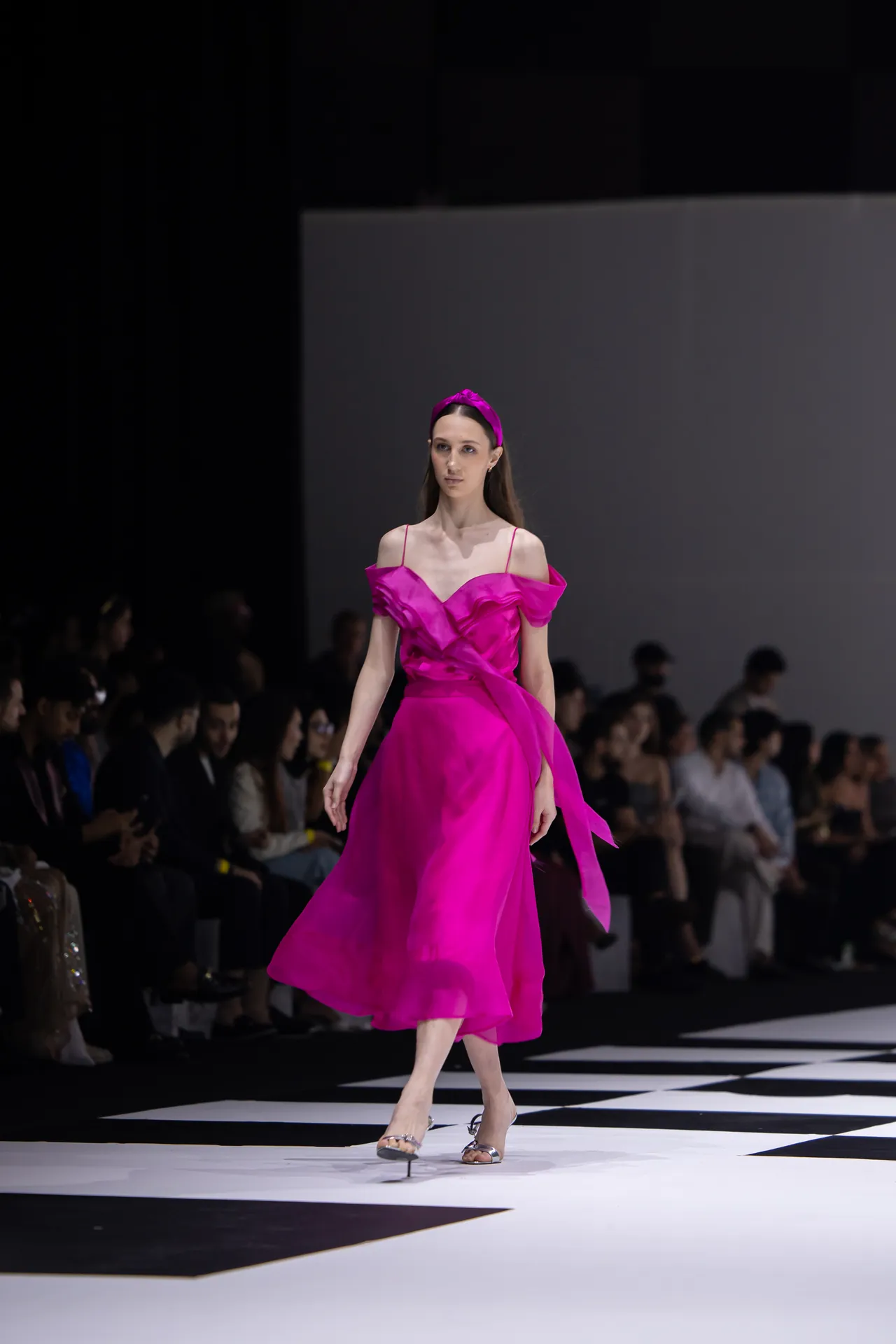 Model walking the runway in a bright pink off-shoulder satin gown with a matching headband, audience in the background.