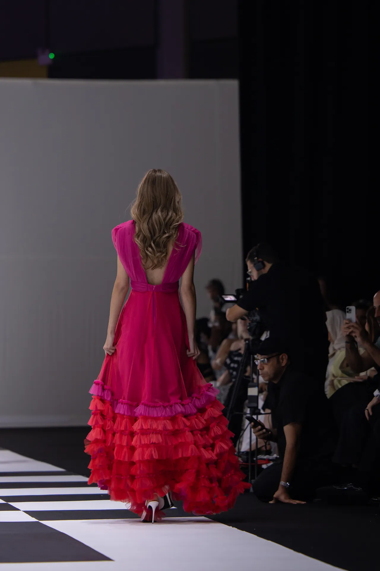 Model walking away on a fashion runway in a bright pink, multi-tiered ruffled gown; photographers and audience line the sides of the catwalk.