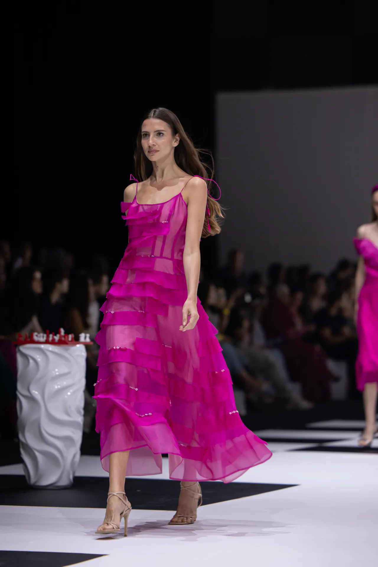 Model walks the runway in a bright pink, tiered sheer dress with beige strappy heels; audience blurred in the background.