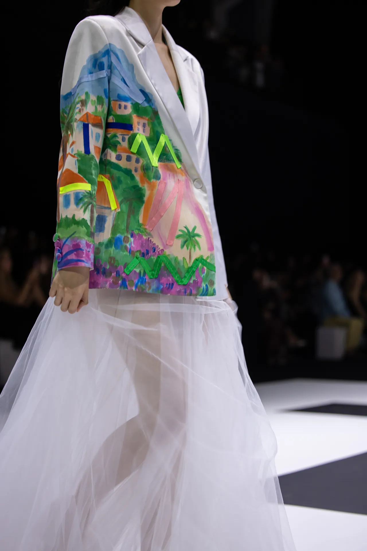 Model wearing a white blazer with a vibrant tropical painting on the side and a sheer white skirt for a fashion show.