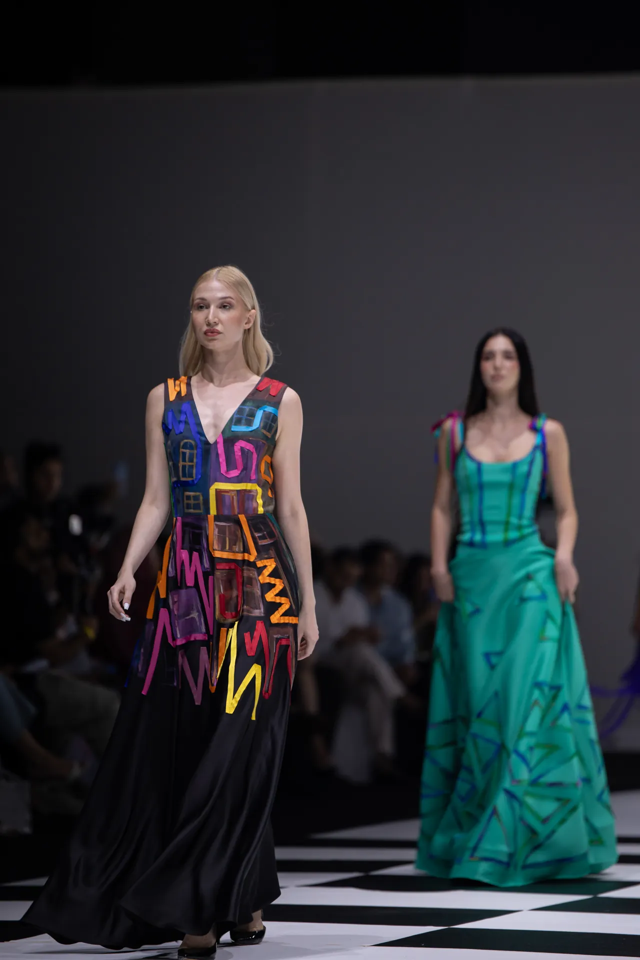 Two models walk a fashion runway; the foreground model wears a sleeveless black gown with bright abstract geometric patterns, while a second model in a turquoise dress stands behind on a checkered runway.