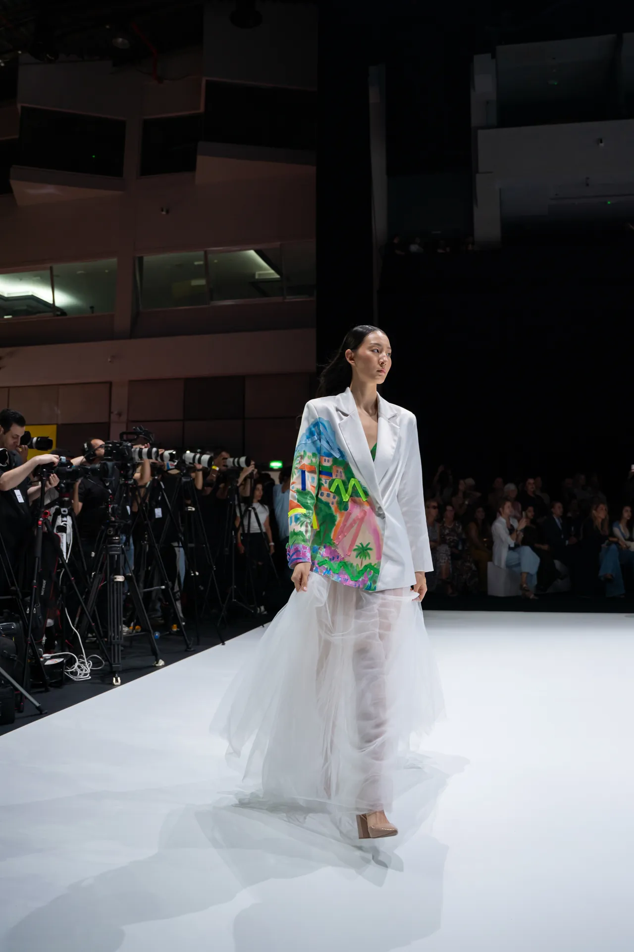 Model walking a white runway in a white blazer with a colorful tropical print and sheer skirt; photographers line the left side of the scene.
