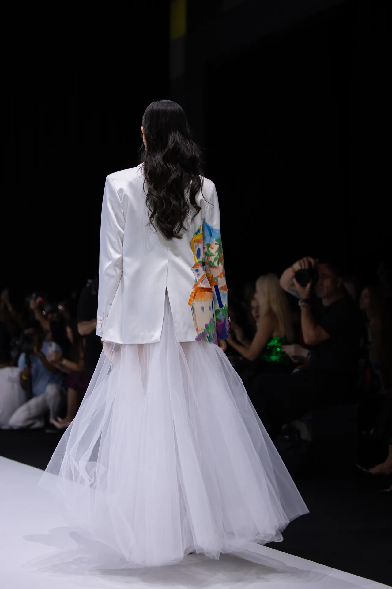 Back view of a model walking the runway in a white blazer with a colorful painted panel and a white tulle skirt.