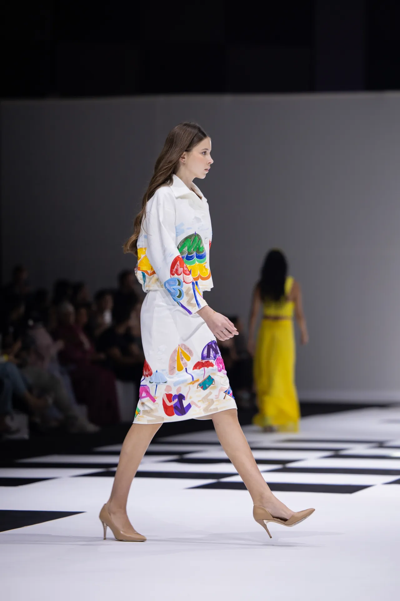 Female model walks a fashion runway in a white dress with colorful abstract prints, beige heels, and a black-and-white checkerboard floor.