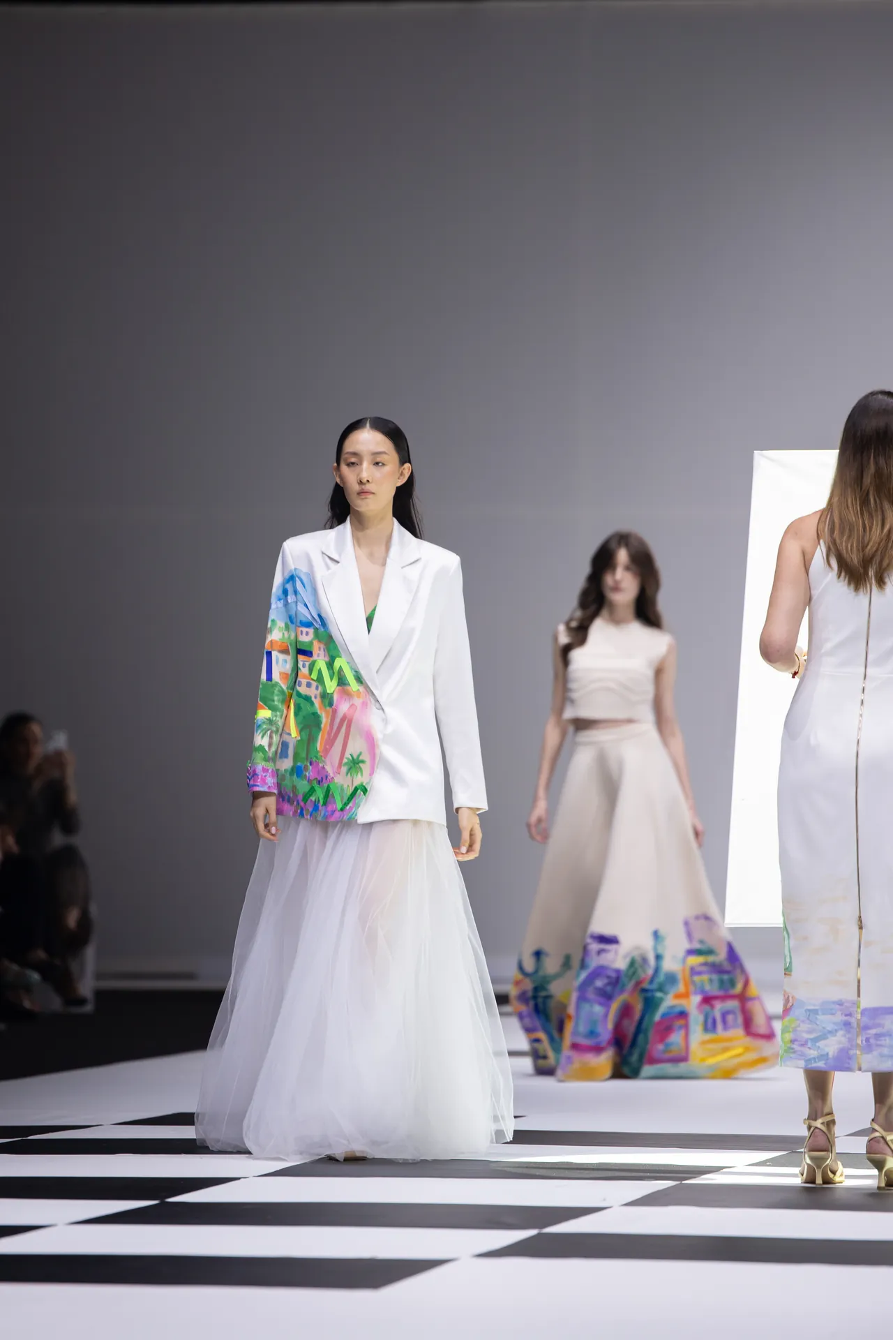 Model walks a fashion runway in a white blazer with a colorful abstract print and a sheer white skirt/veil.