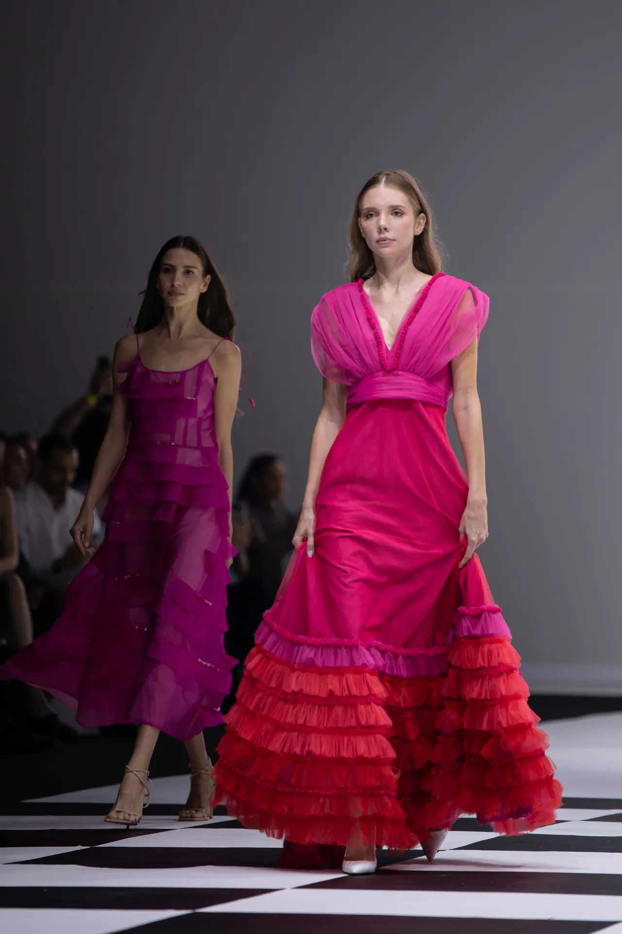Model in a bright pink, ruffled gown walks the runway as another model in a pink dress follows behind on a black-and-white checkered catwalk.