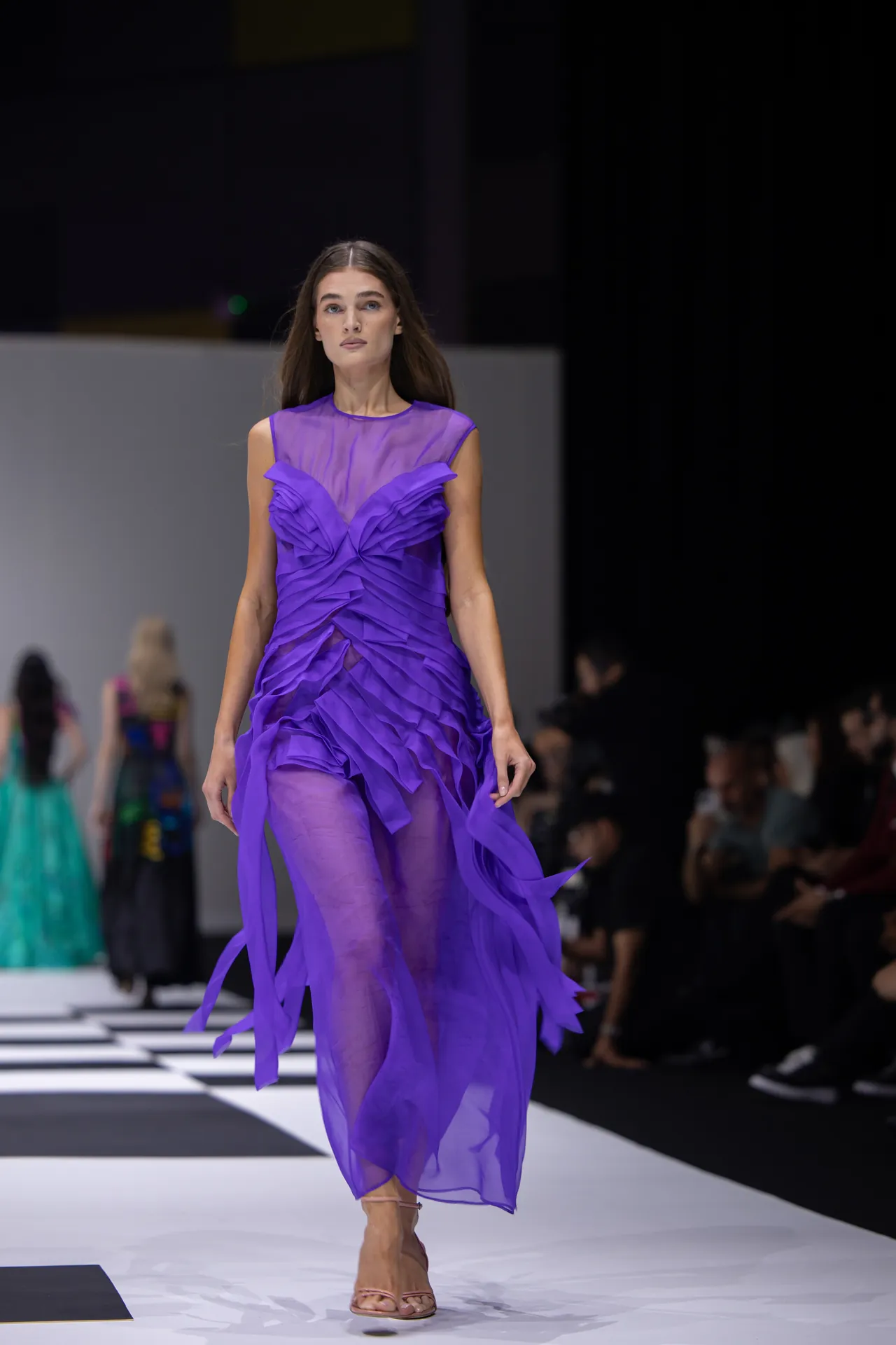 Runway model walking in a sheer purple gown with layered ruffles and beige strappy sandals.