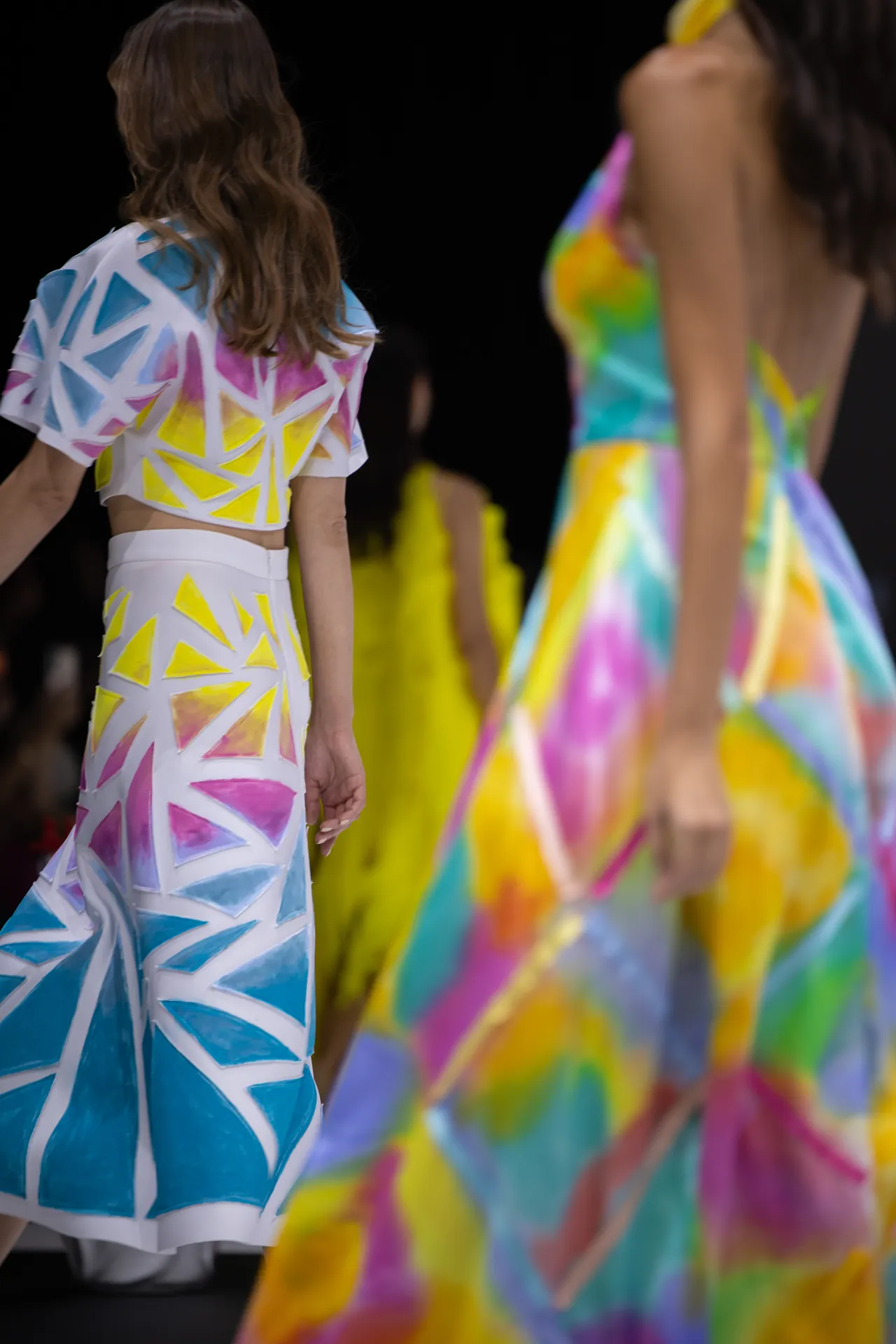 Models walk the runway in bold, geometric print dresses featuring blue, yellow, pink triangles on white fabrics. The foreground model wears a cropped top and midi skirt; colorful gowns trail in the background.