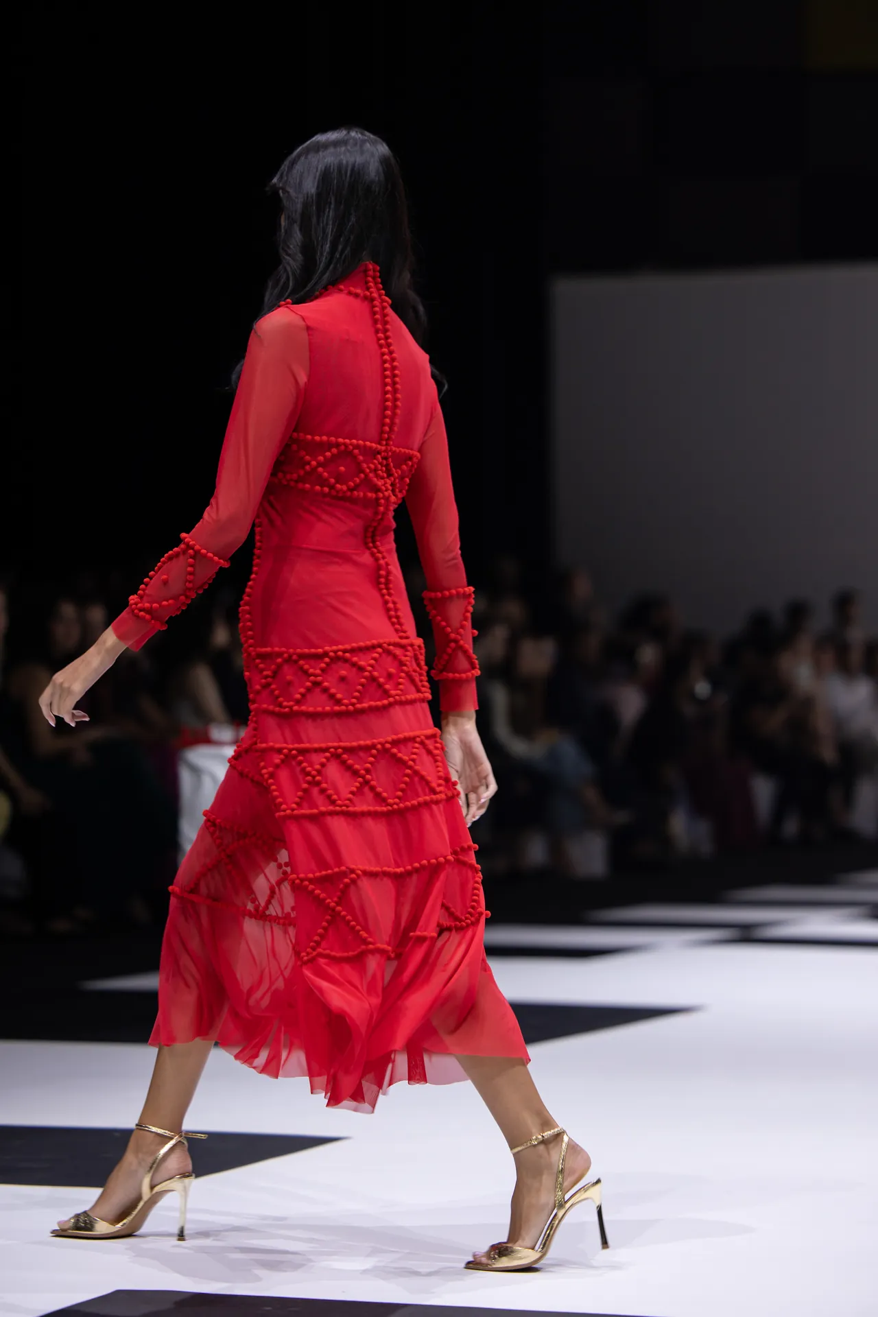 Runway model walking away in a red, beaded dress with geometric beadwork, gold heels, and an audience in the background.