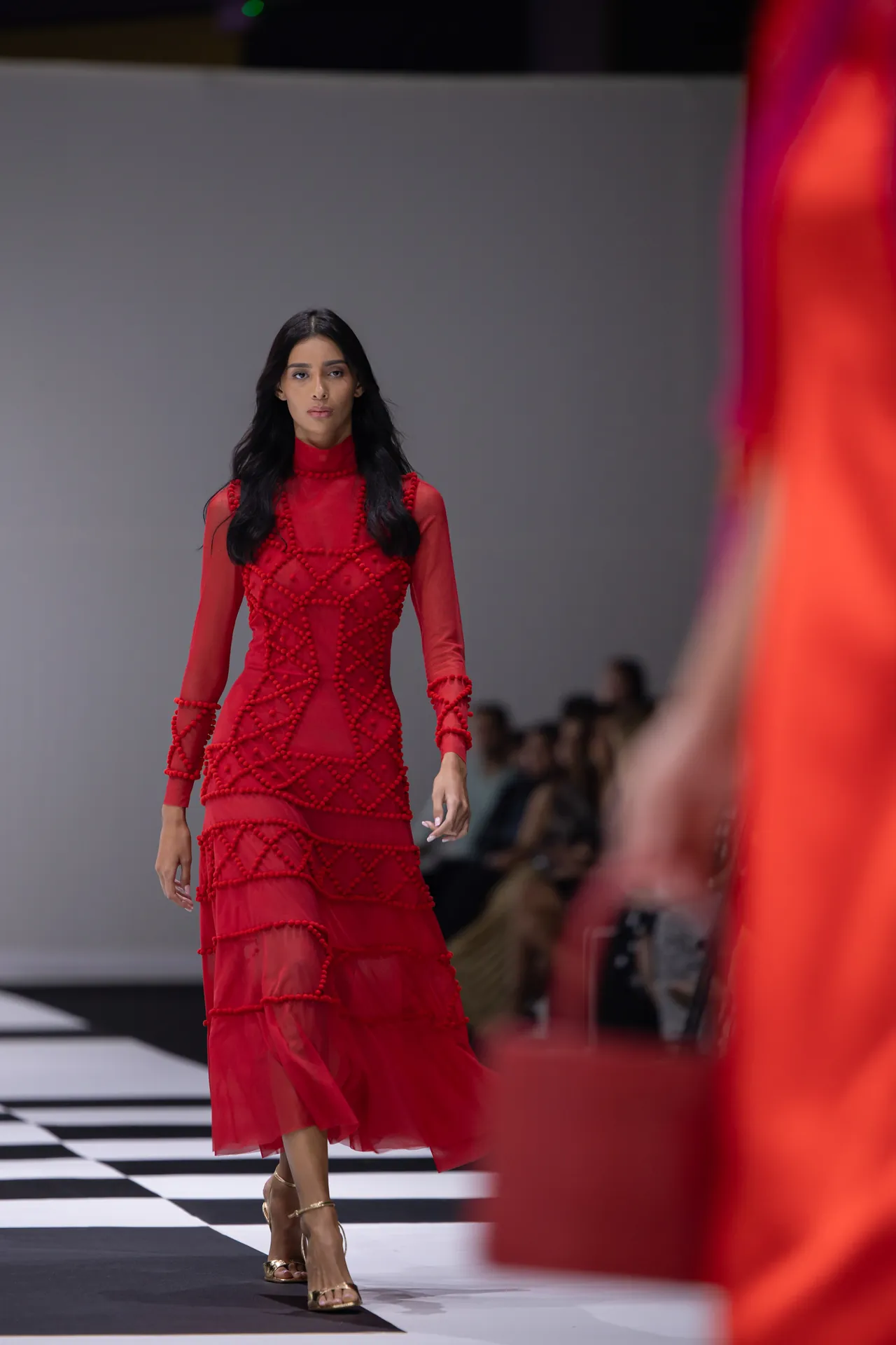 Model walking the runway in a sheer red gown with geometric beadwork, on a black-and-white checkerboard floor.