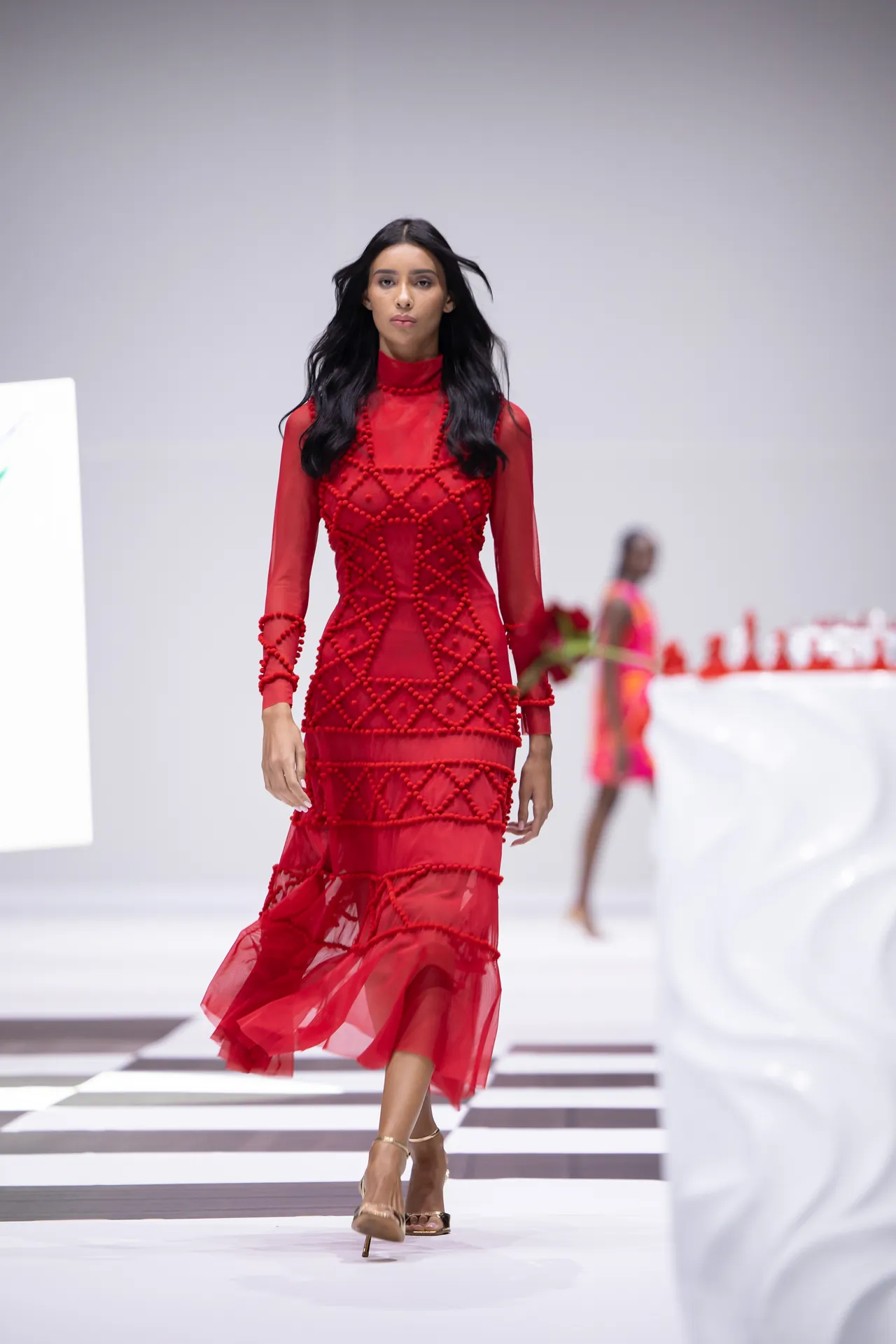 Model walks a runway in a red sheer gown with bead lattice detailing and high-heeled sandals.