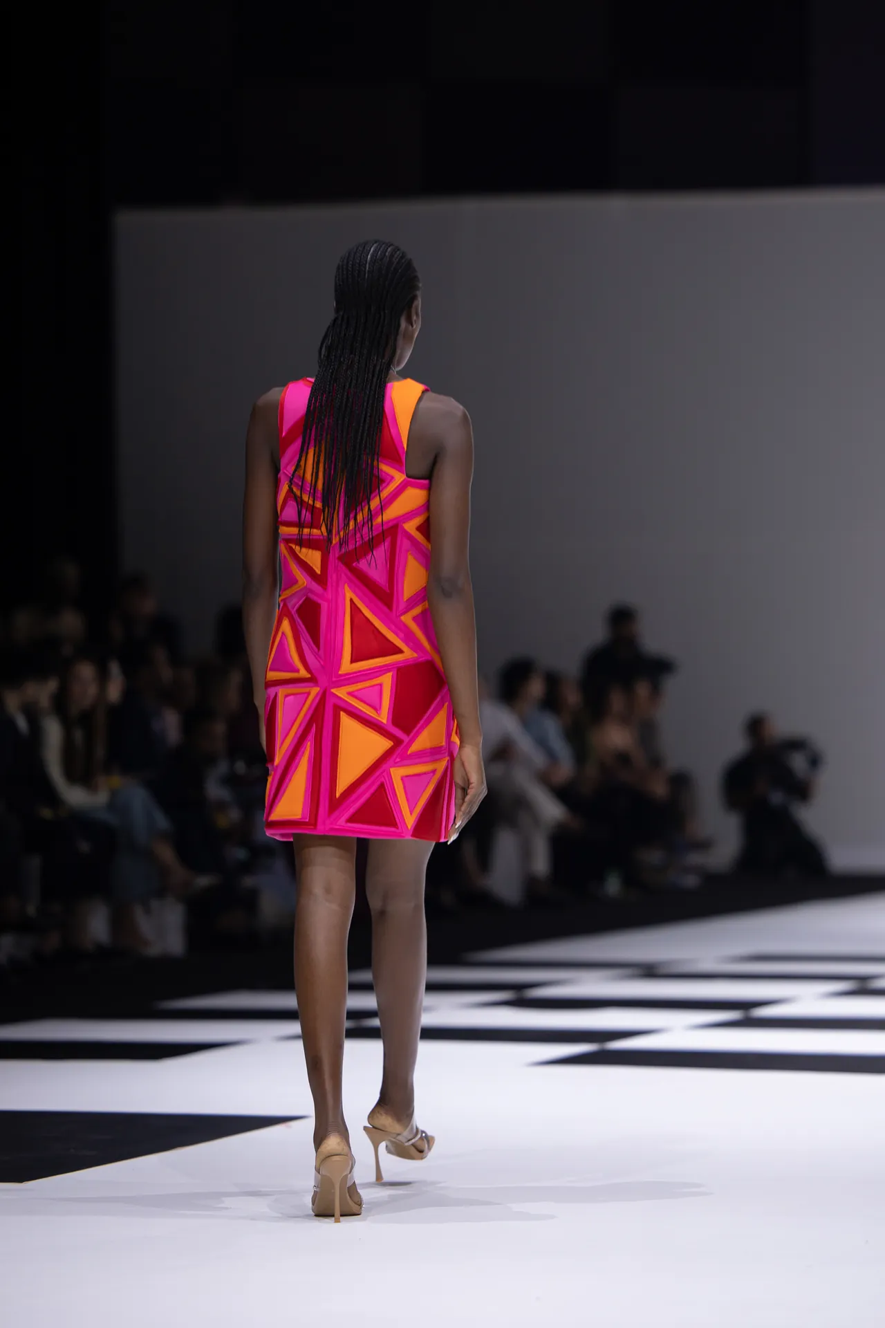 Model seen from behind walking a runway in a neon pink and orange geometric-patterned sleeveless dress, beige heels.