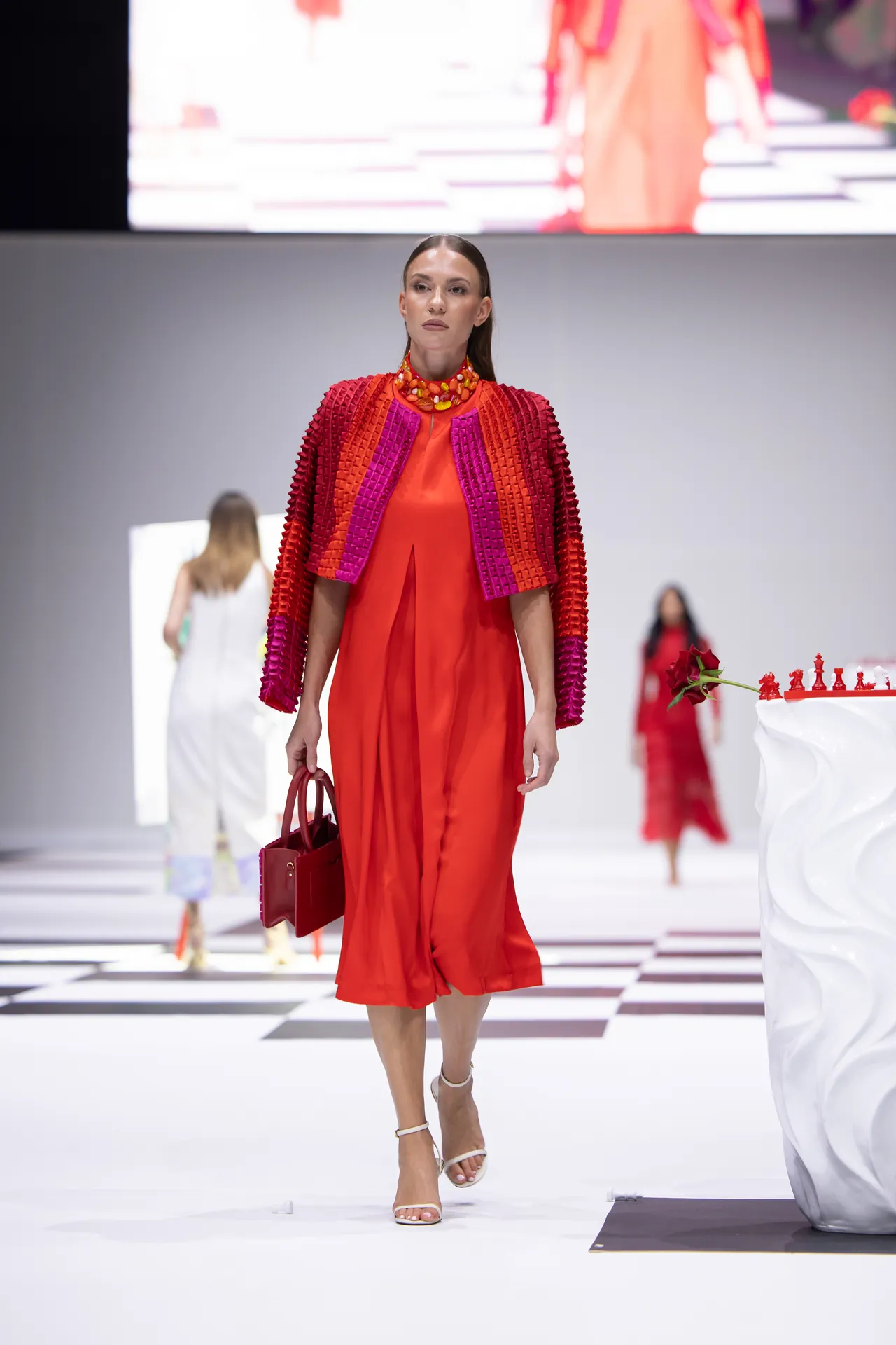 Model walks a fashion runway in a bright red dress with a textured pink-red cape, carrying a red handbag and wearing a chunky orange necklace.