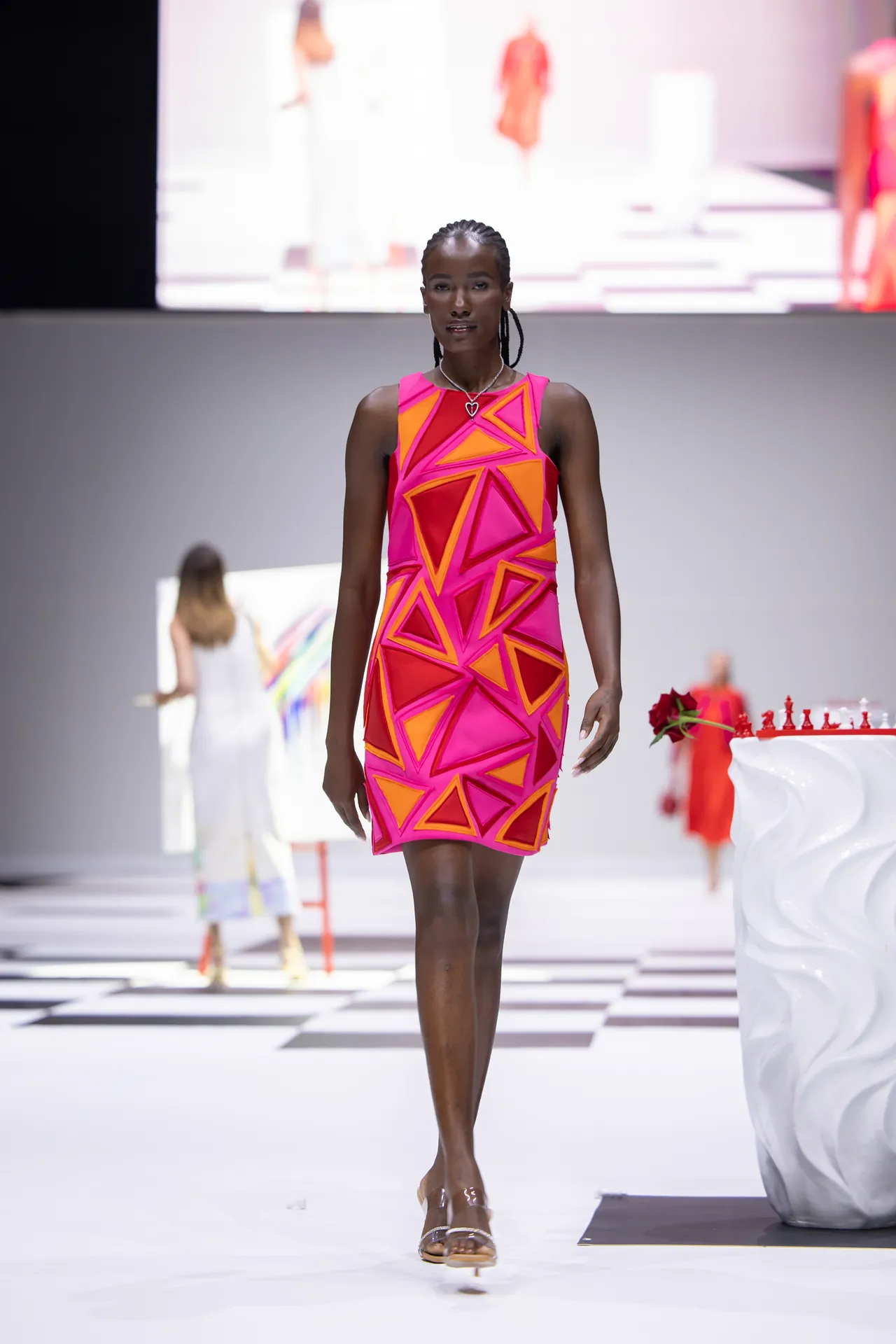 Model walks the runway wearing a bright pink dress with orange geometric triangles in a fashion show.
