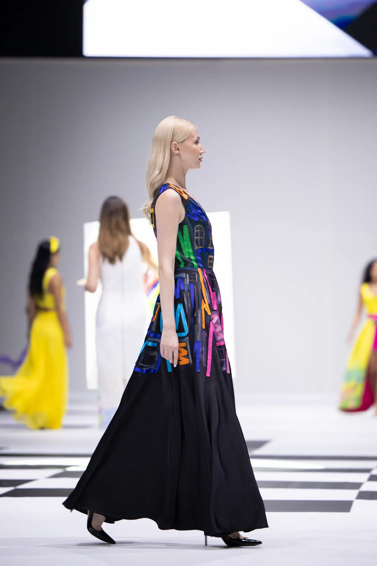 Side-profile of a fashion model walking the runway in a long, colorful dress with a black hem, on a checkered catwalk.