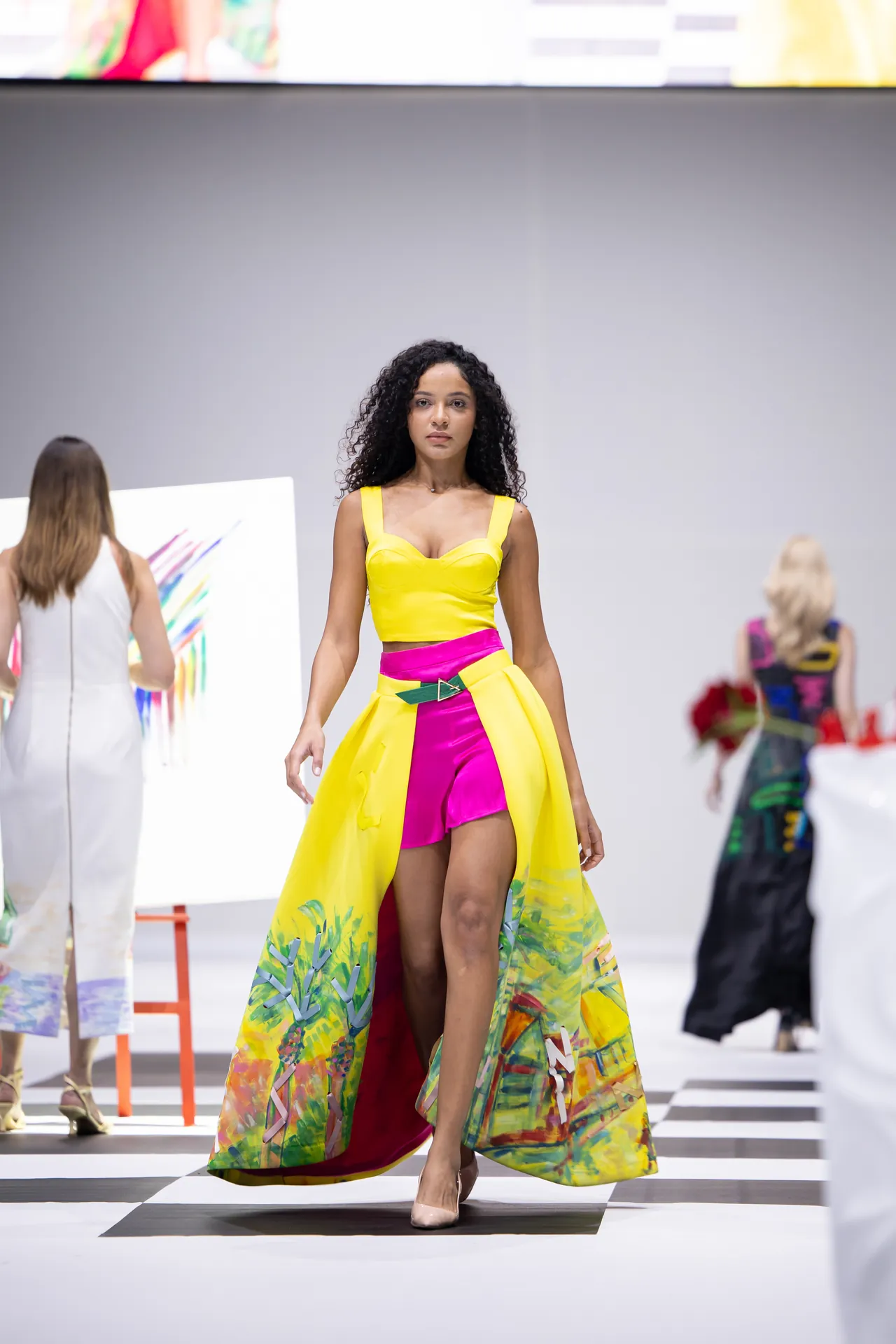 Model walks the runway in a bright yellow crop top and vibrant color-block cape with pink shorts, tropical print flowing behind her.