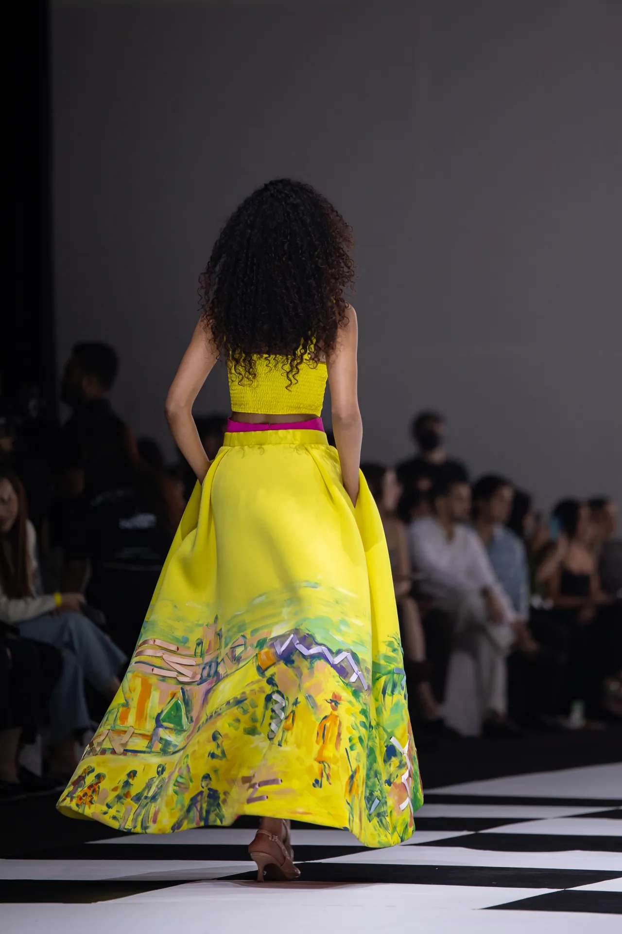 Model walking away on a runway in a bright yellow crop top and a long, colorful abstract skirt; audience blurred in the background.