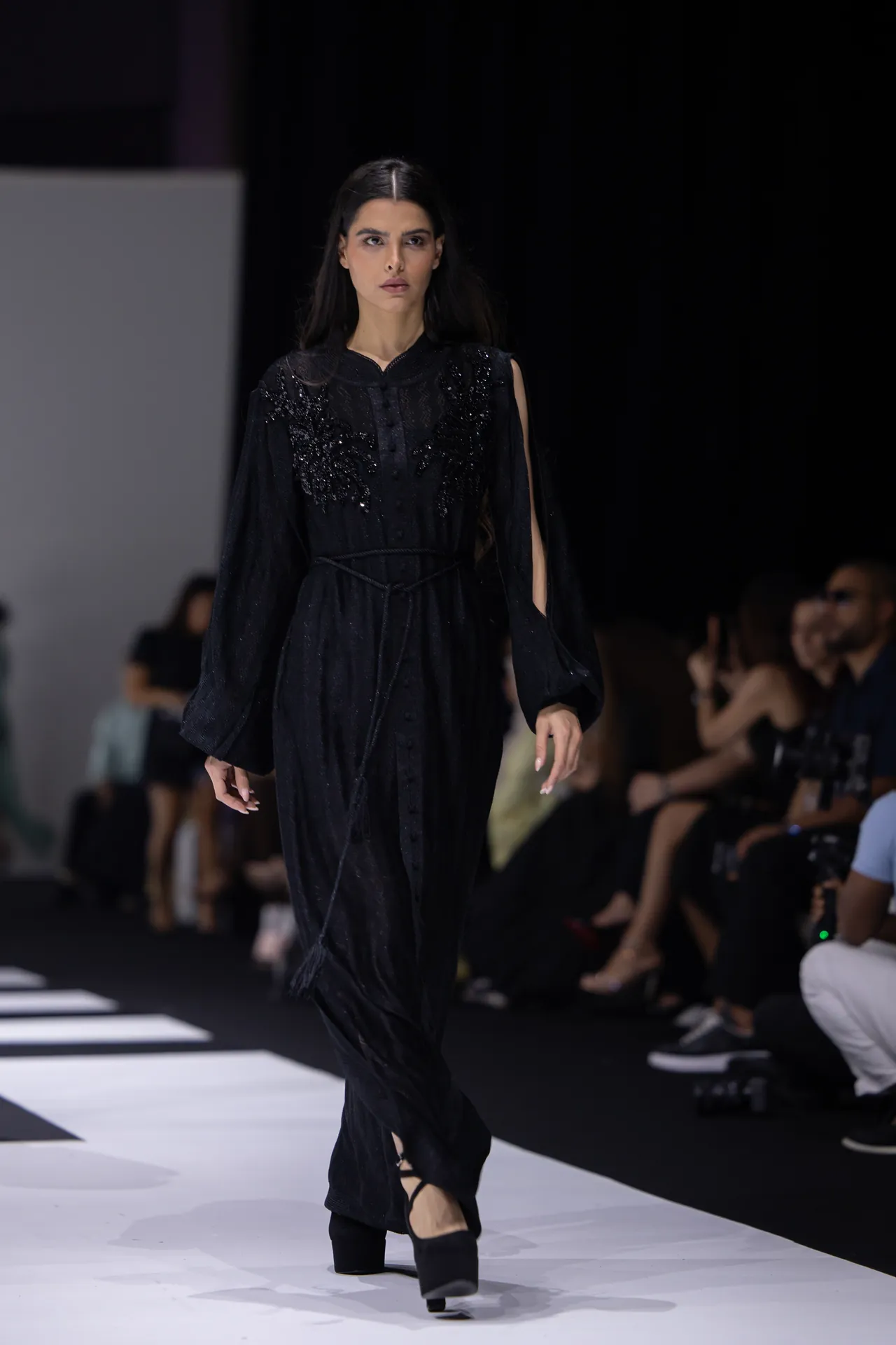 Female model walks a black, embellished gown down a fashion runway; audience seated along the sides.