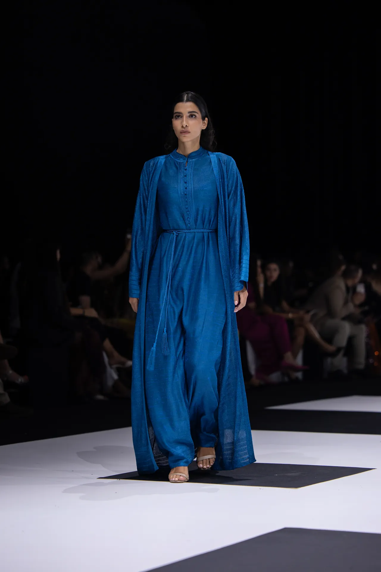 Model walking a runway in a blue, floor-length dress with a matching belt; dark audience background remains out of focus.