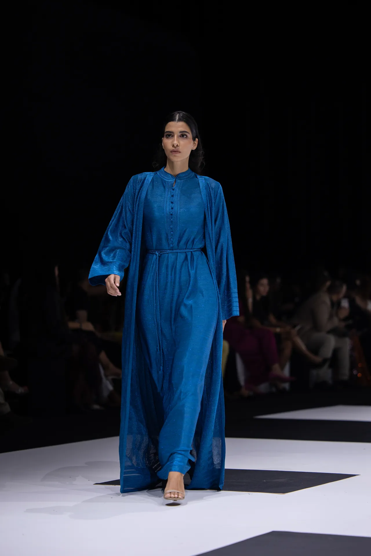 Model walking a fashion runway in a flowing royal blue dress with matching cape, barefoot on a white and black patterned catwalk.