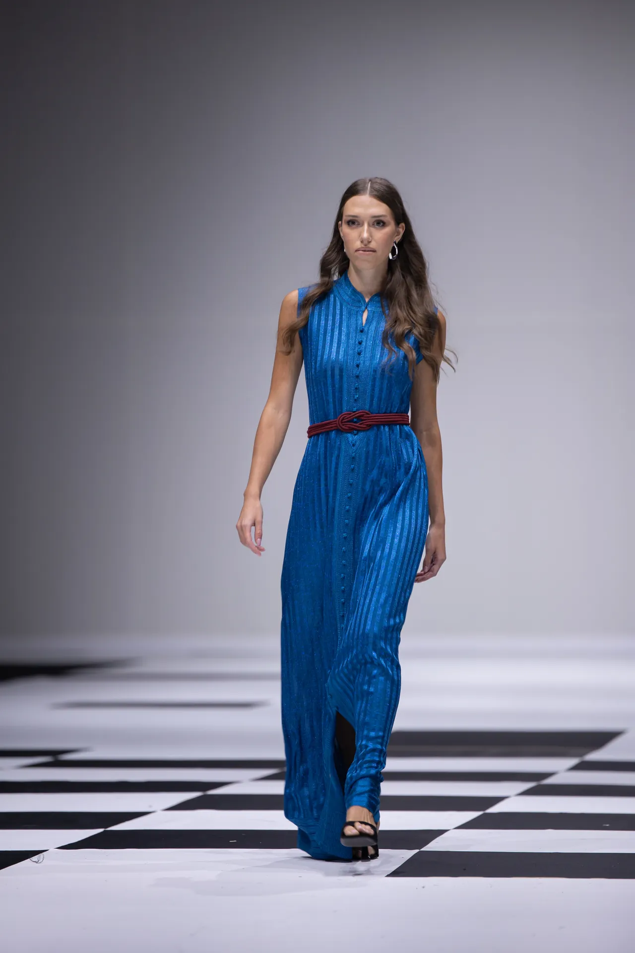 Female model walking the runway in a blue striped sleeveless dress with a red belt, on a black-and-white checkered floor.