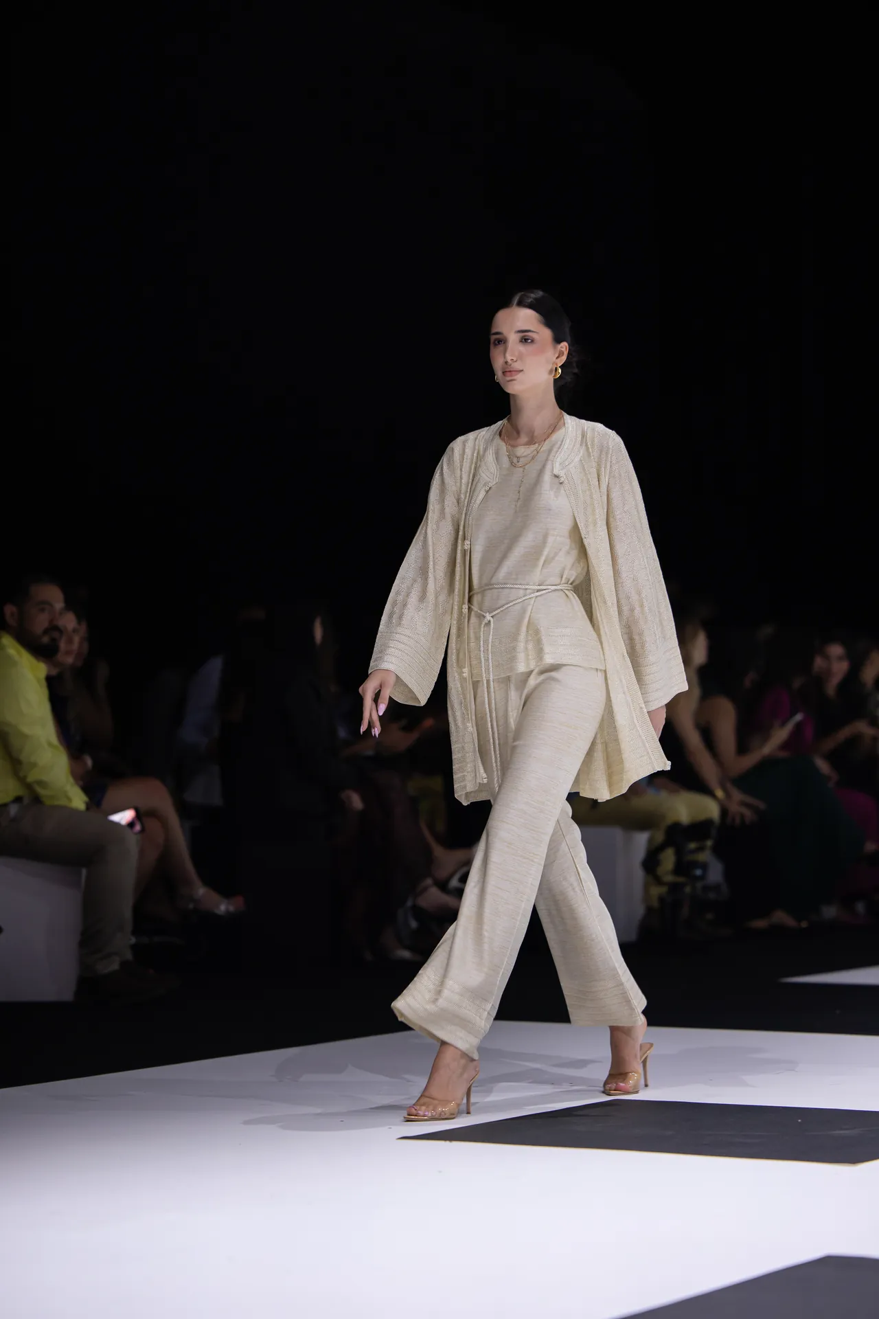 Model walking the runway in a cream textured suit with a matching long cardigan and belt, beige heels visible against a dark backdrop