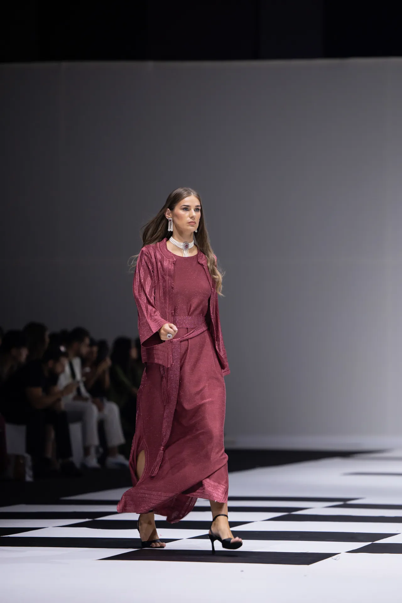 Model walks a fashion runway wearing a burgundy matching set with a sheer outer layer and statement jewelry.