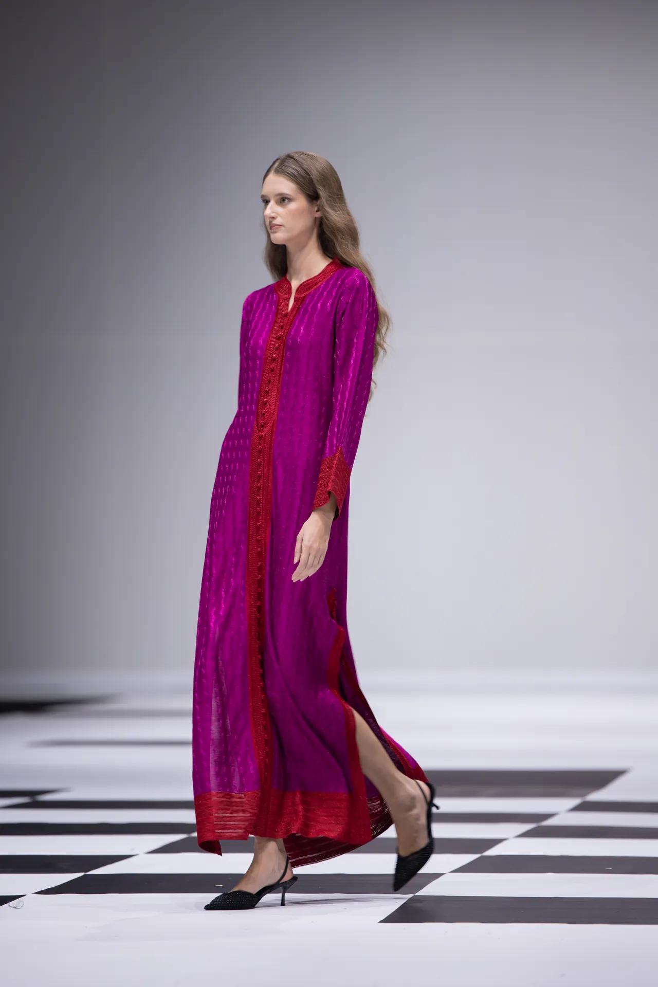 Model walking the runway in a bright magenta dress with red trim, on a black-and-white checkerboard runway.