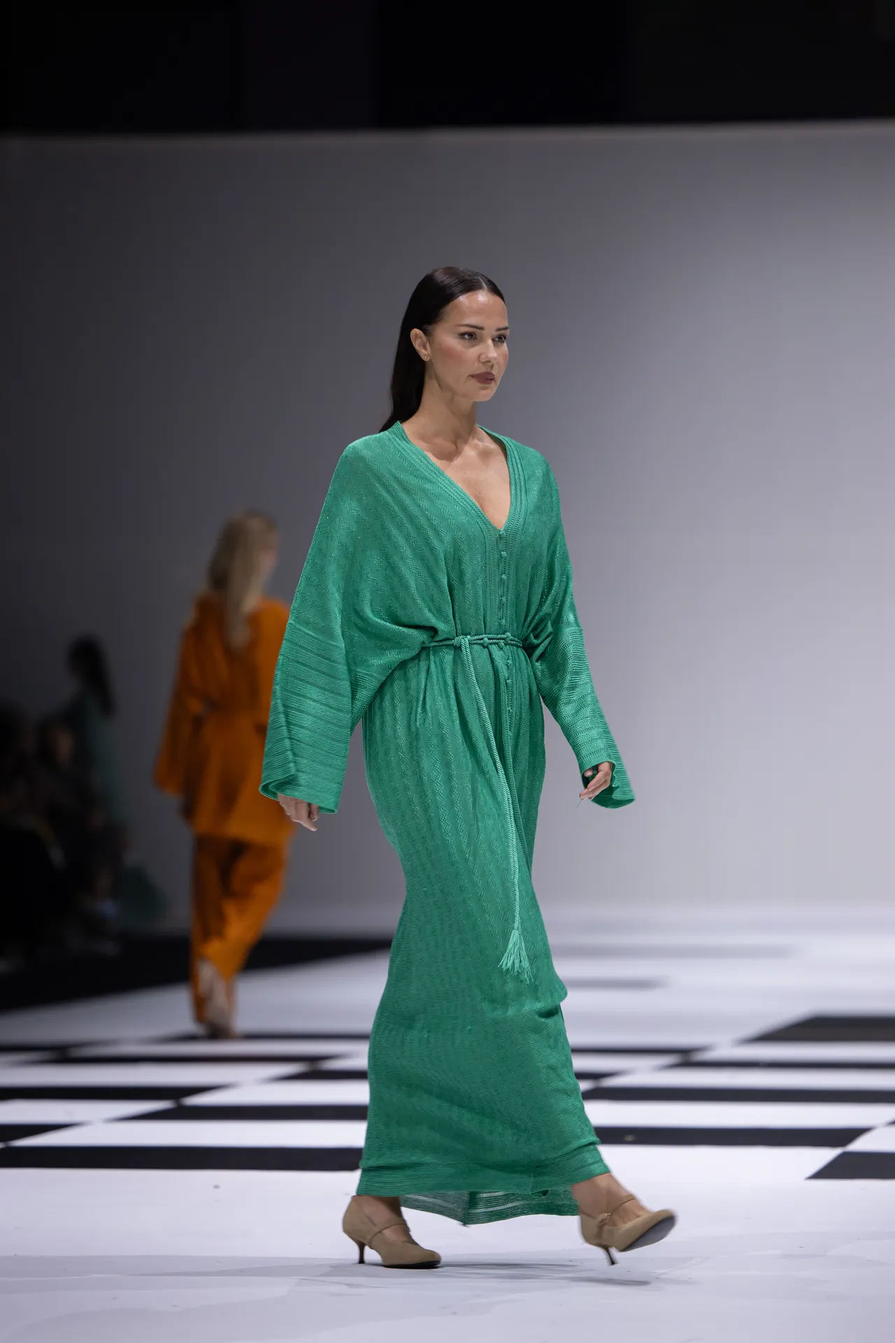 Model walks the runway in a green textured wrap dress with a tied waist and long sleeves, beige heels, on a black-and-white checkered floor.