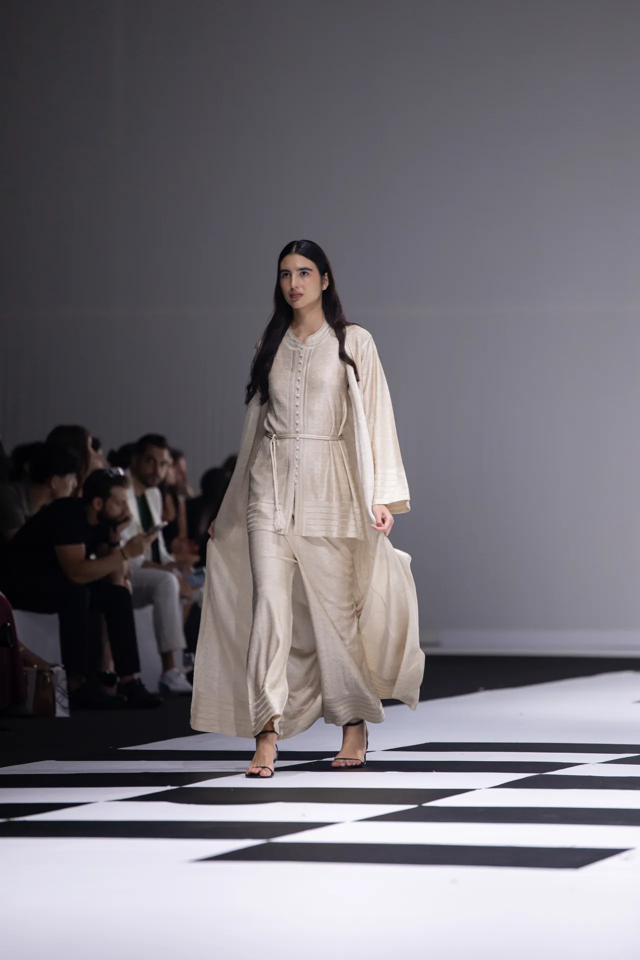 Female model walks a beige, flowing outfit down a fashion-show runway.