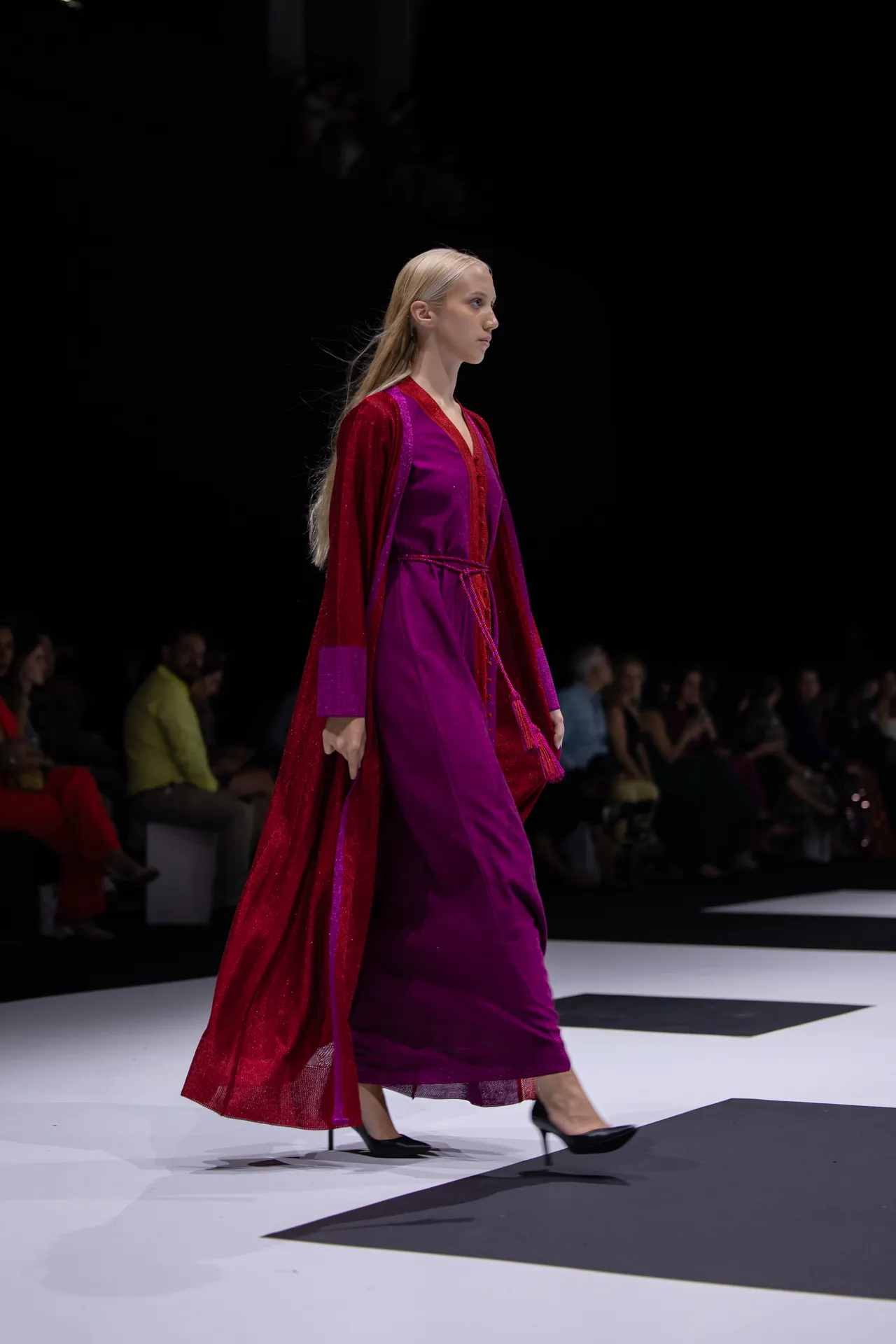 Model walking the runway in a magenta dress with a red cape at a fashion show, audience visible in the background beside a white catwalk.