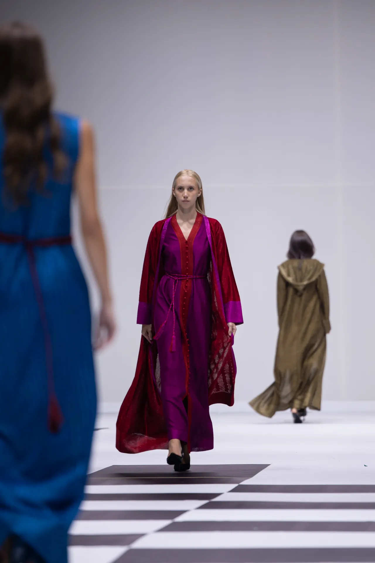 Front-and-center model in a vivid magenta gown with a cape walks the runway, others in blue and tan outfits blur in the background.