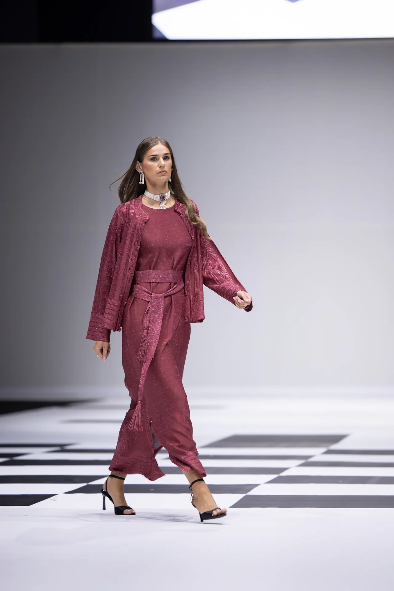 Model walking the runway in a burgundy textured suit with a matching jacket and tassel belt on a black-and-white checkered catwalk.