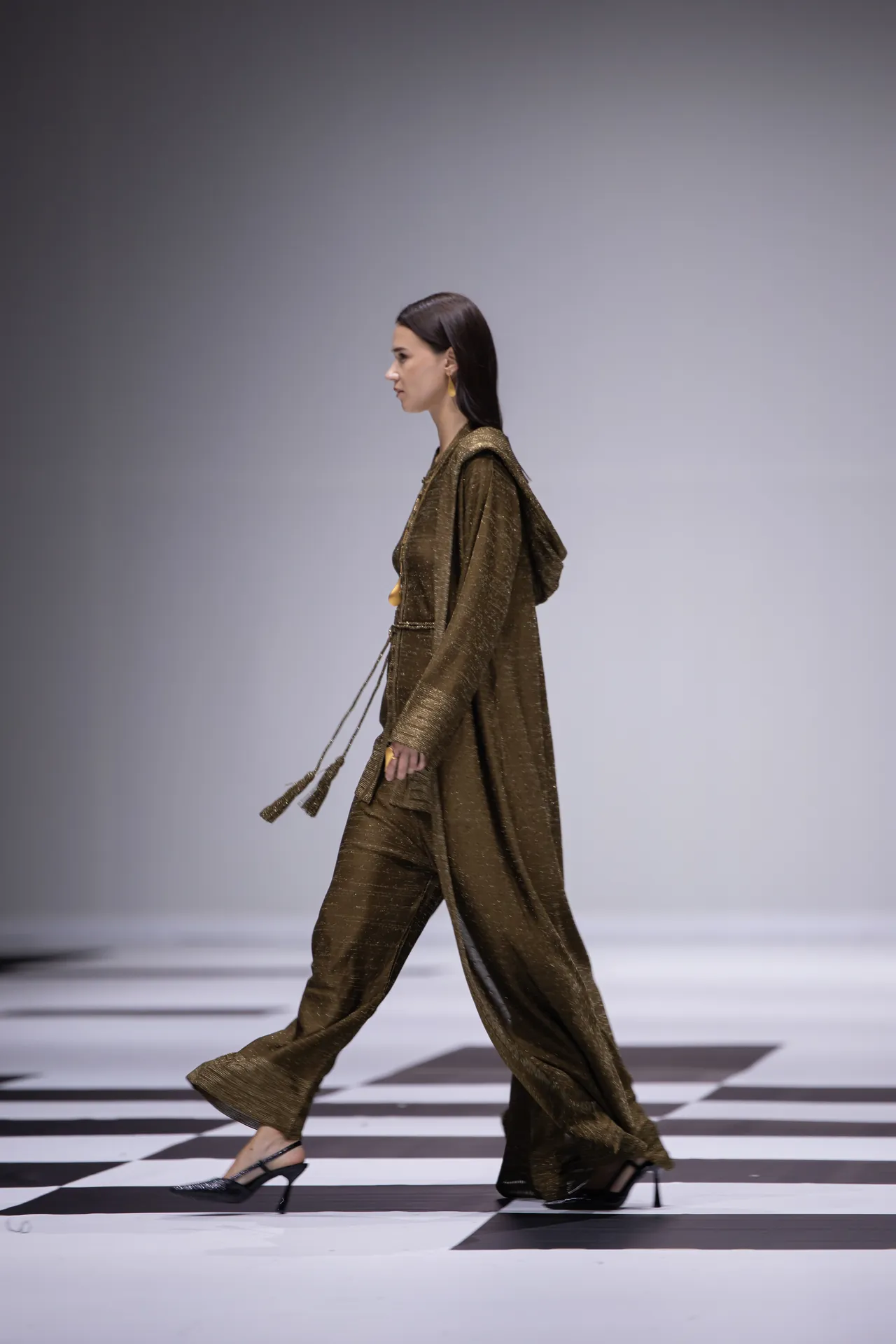 Female model walking a fashion runway in a shimmering bronze-brown beltable coat and matching pants, paired with black heels.