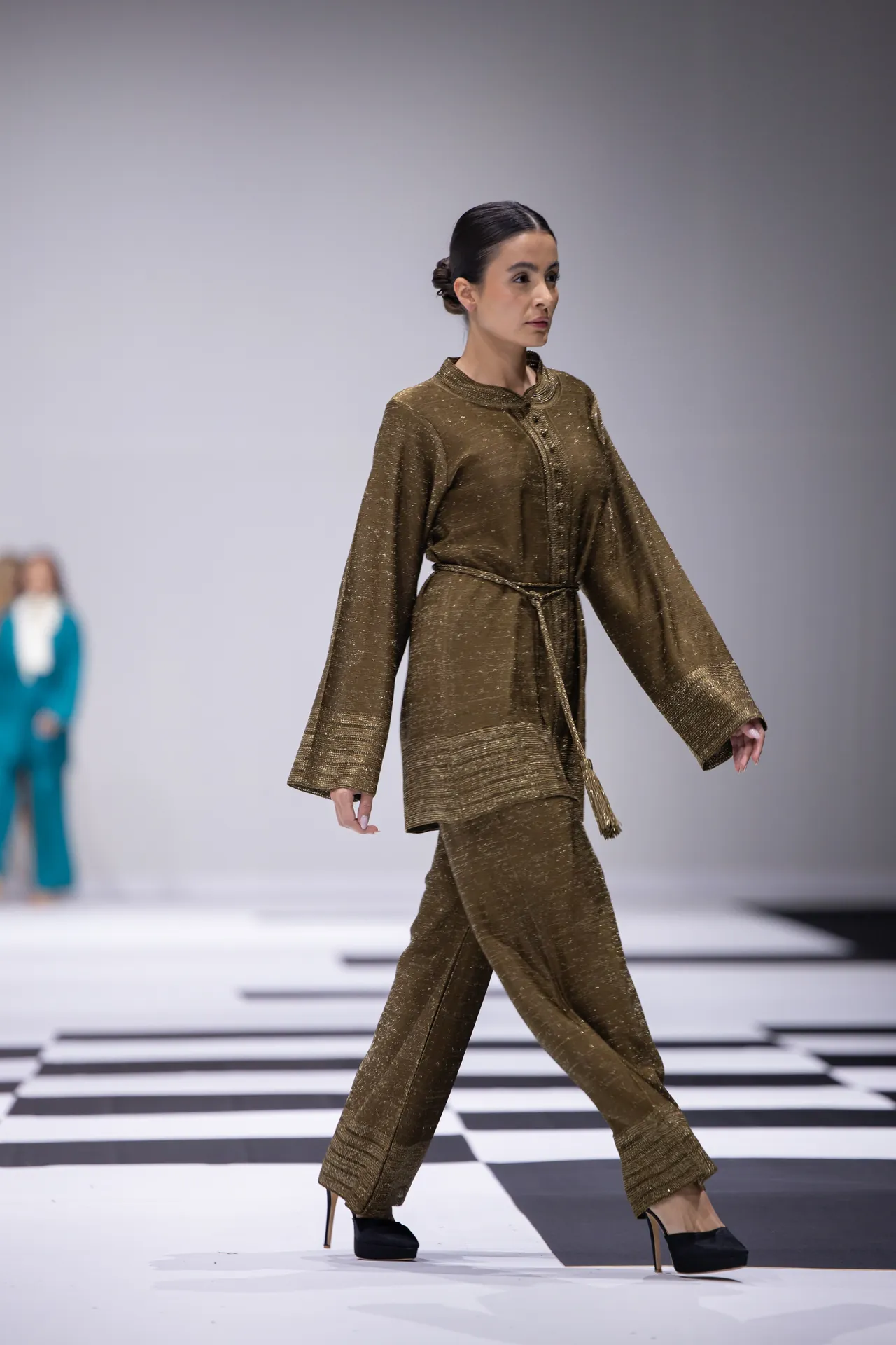 Model walks a fashion runway wearing a gold metallic matching set with a belt and black heels on a black-and-white checkered catwalk.”] ,