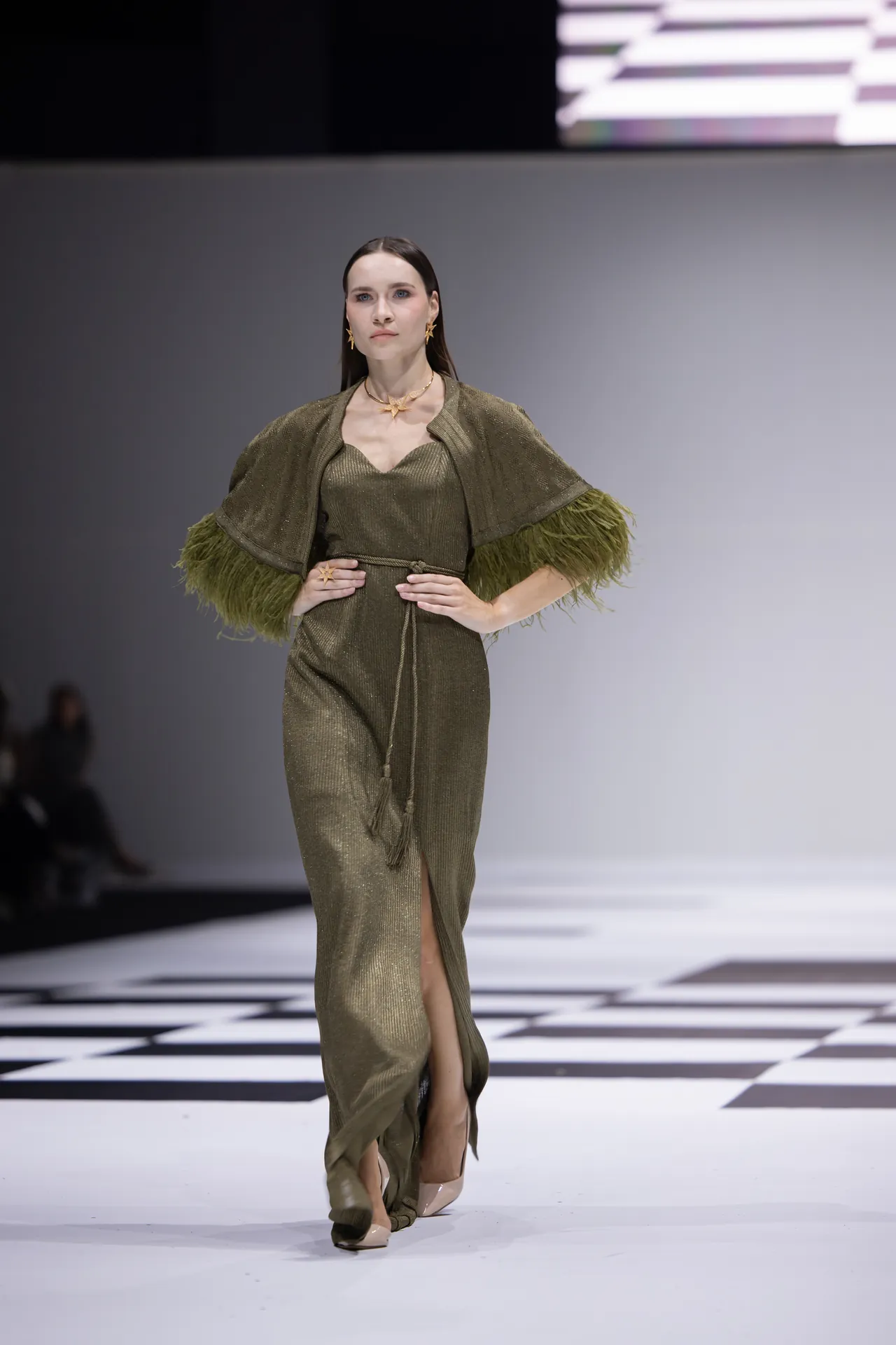 Model walks the runway in a metallic olive-green dress with feathered sleeves and a waist belt on a checkered catwalk.