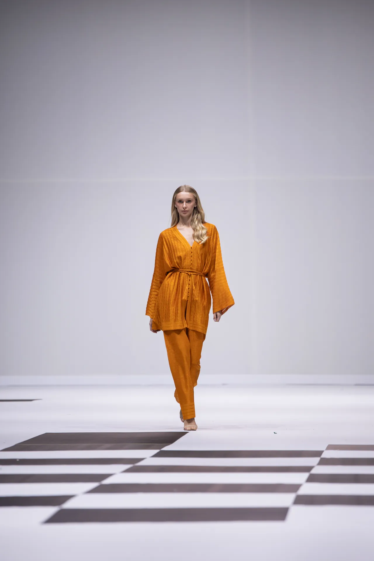 Model walking the runway in an orange belted outfit on a minimalist white stage with a black-and-white checkered floor pattern.